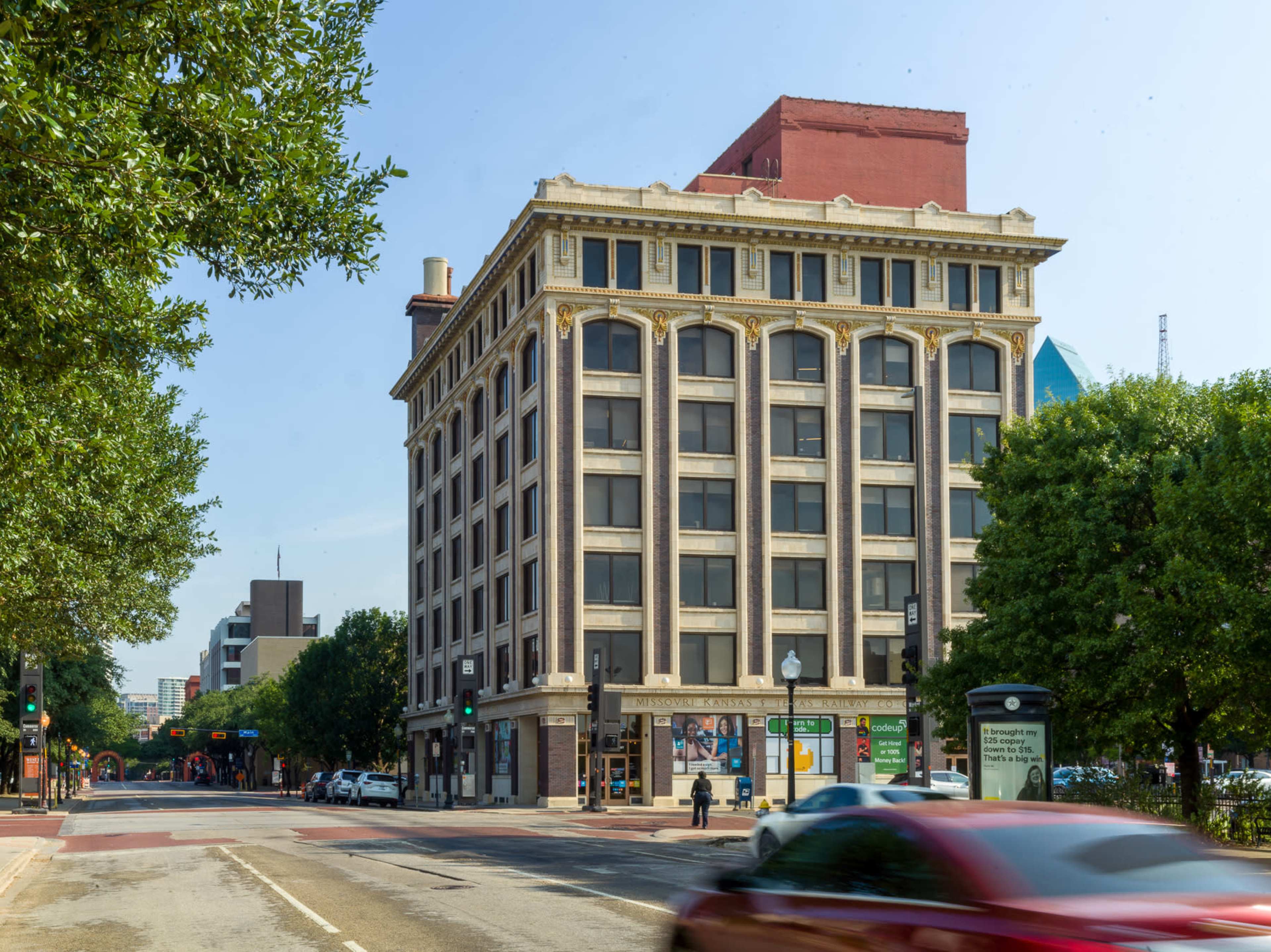 The image shows a multi-story building with a decorative facade located at the corner of a city street, surrounded by trees and parked cars.