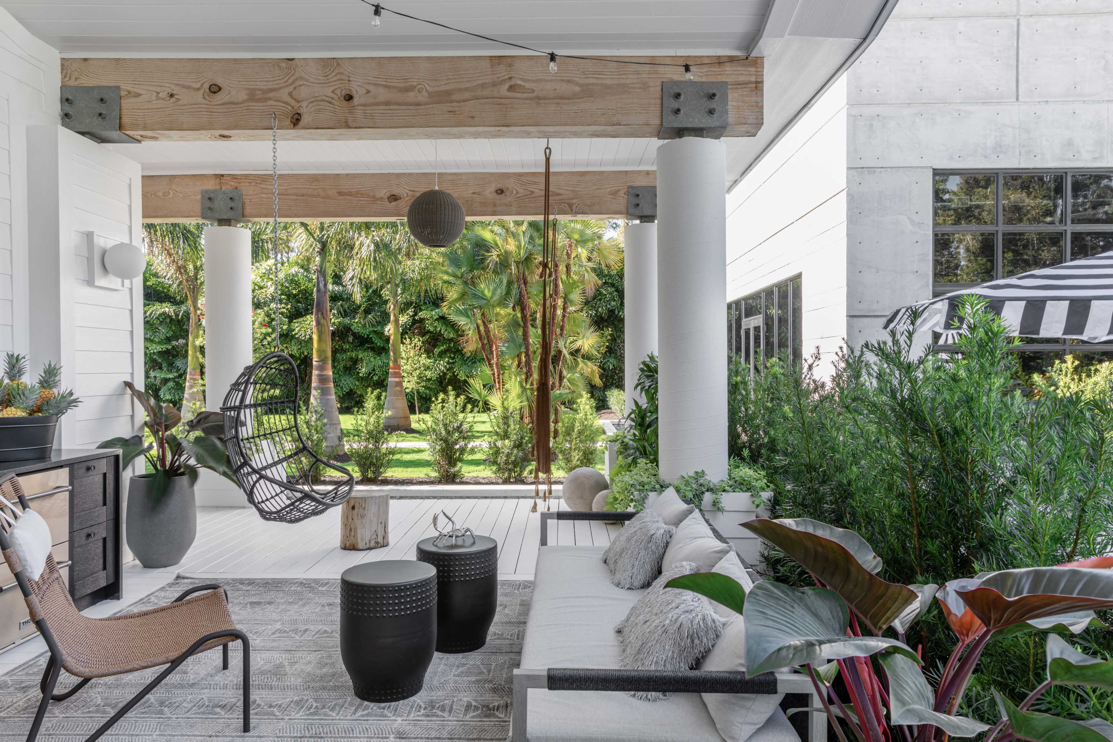 The image shows a modern outdoor patio with a hanging chair, two circular tables, and various plants, set against a backdrop of greenery and a structured building.