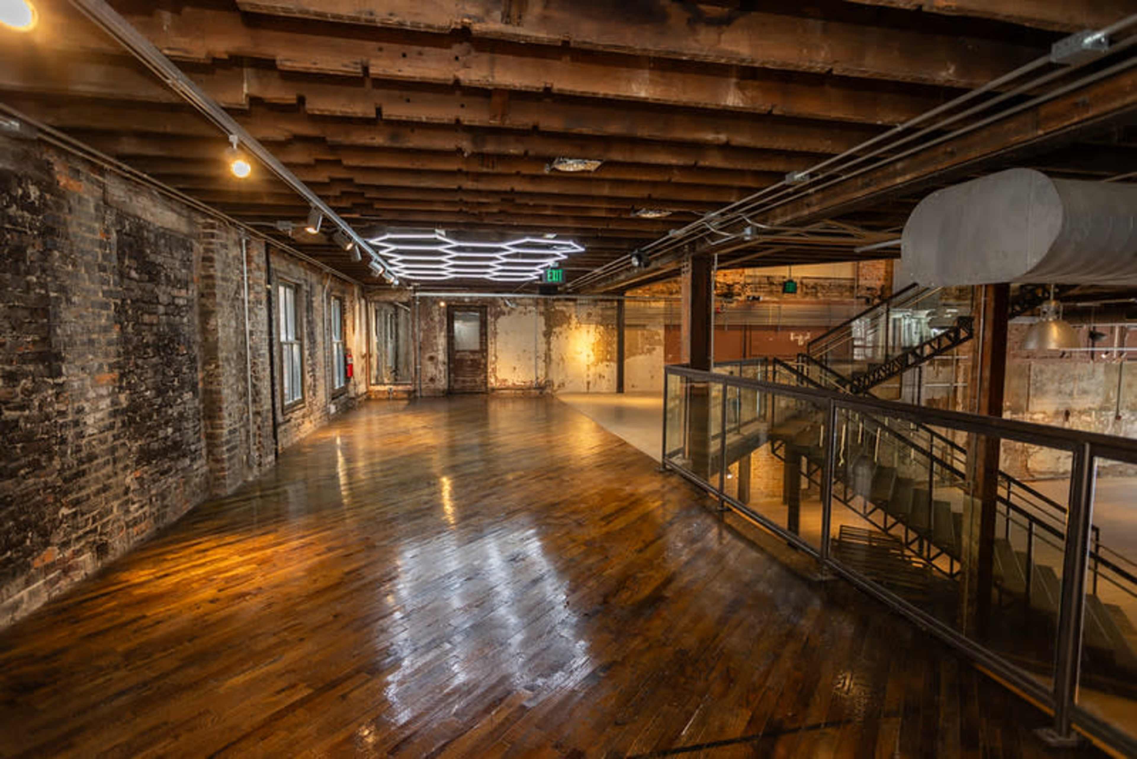 The image shows a spacious, industrial-style interior with exposed brick walls, wooden flooring, and a modern lighting fixture overhead.