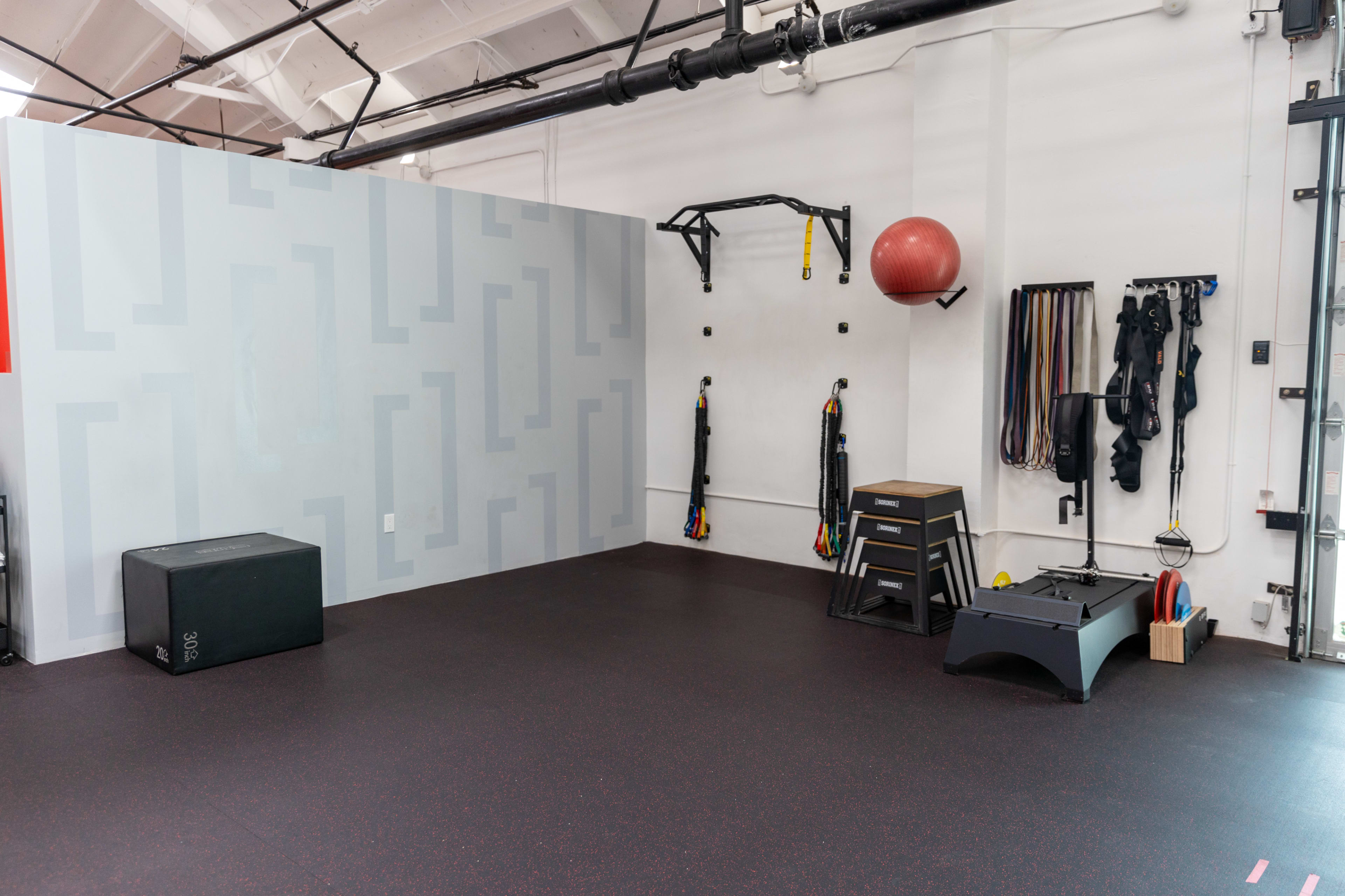 The image shows a gym interior featuring a gray wall with geometric patterns, a black box for workouts, a red exercise ball, and various resistance bands hanging on hooks.