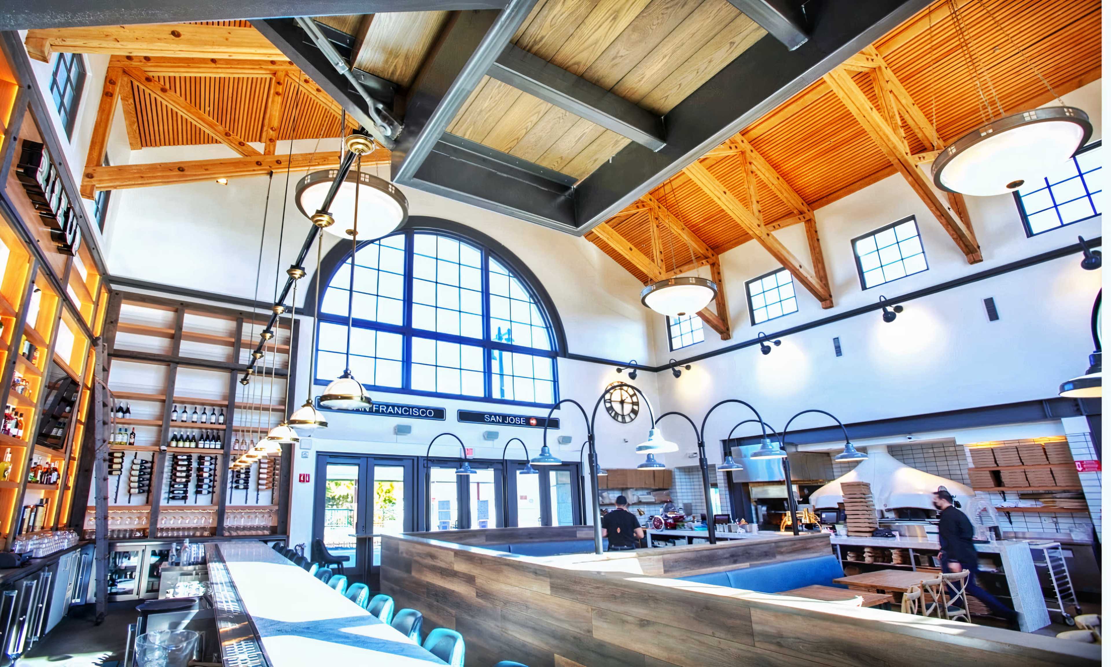 A spacious restaurant interior with a high wooden ceiling, large arched windows, and a long bar area with stools.