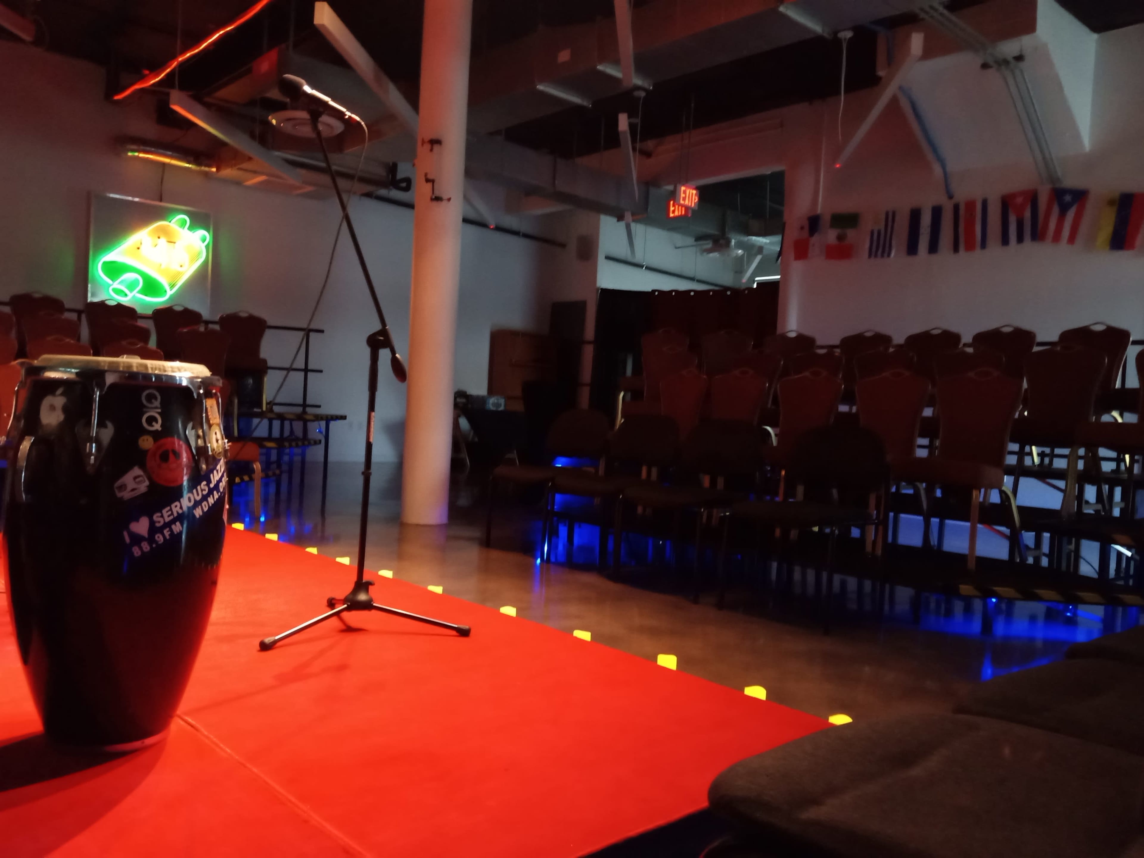 A small performance space featuring a conga drum and a microphone on a red stage, with rows of empty chairs and various international flags hanging on the walls.