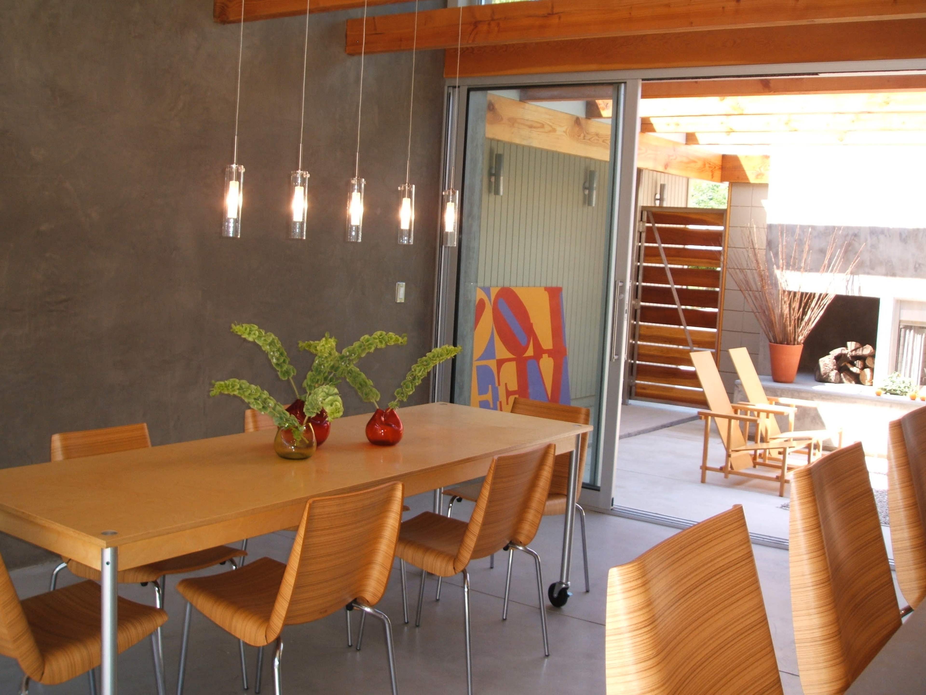 A modern dining area features a long wooden table surrounded by sleek chairs, with glass pendant lights hanging above and large sliding doors leading to an outdoor space.