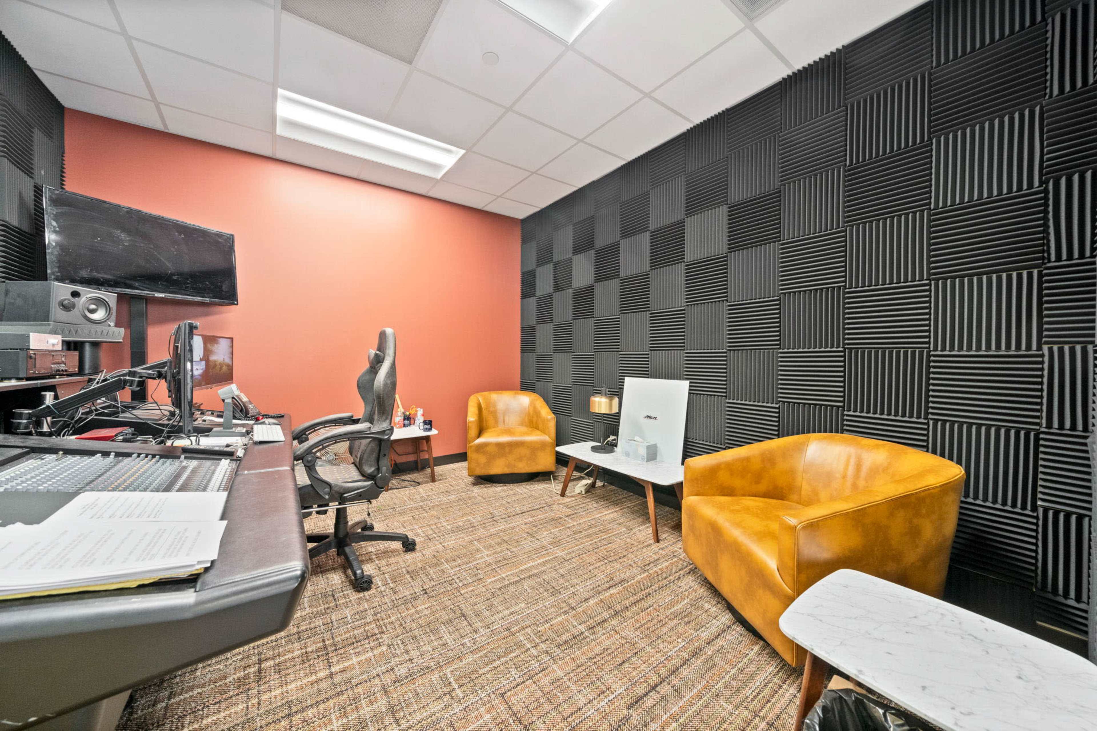 The image shows a sound studio featuring a mixing desk, a large monitor, two brown leather chairs, and soundproof foam panels on the walls.