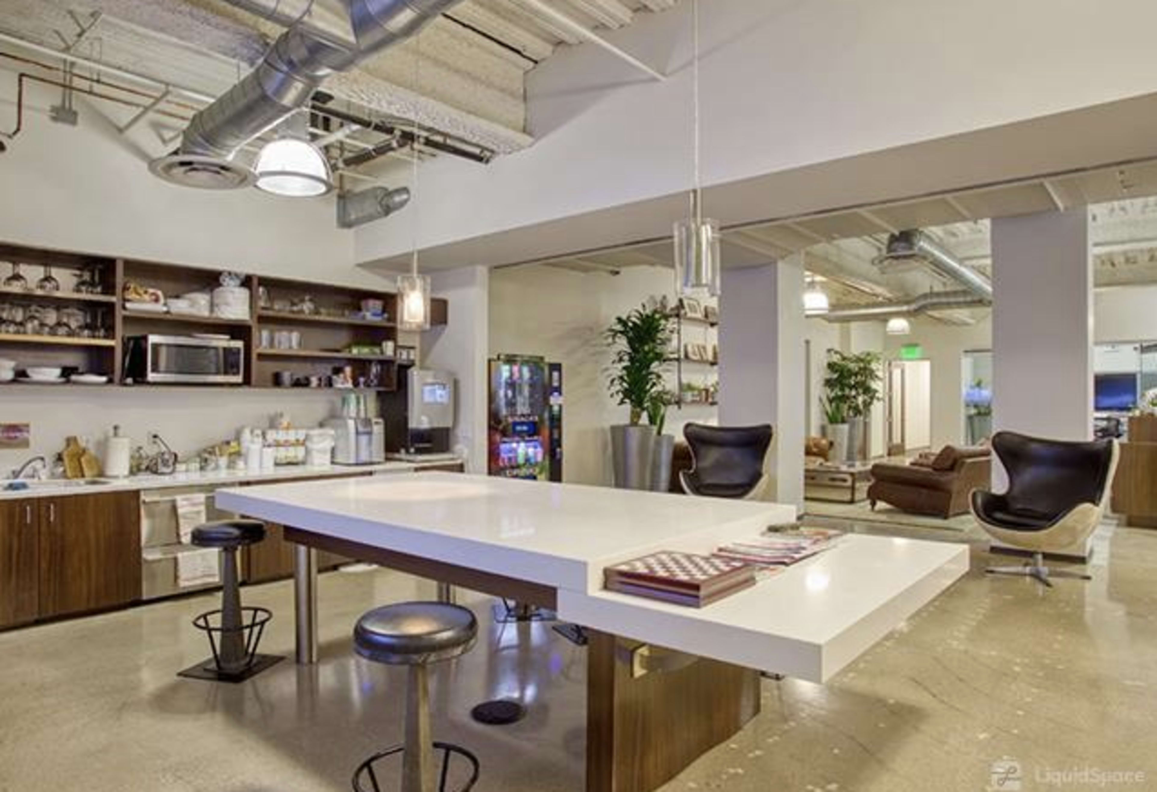 The image shows a modern kitchen area with a long white countertop, wooden cabinets, a kitchenette with a microwave, and seating near a vending machine in an office space.