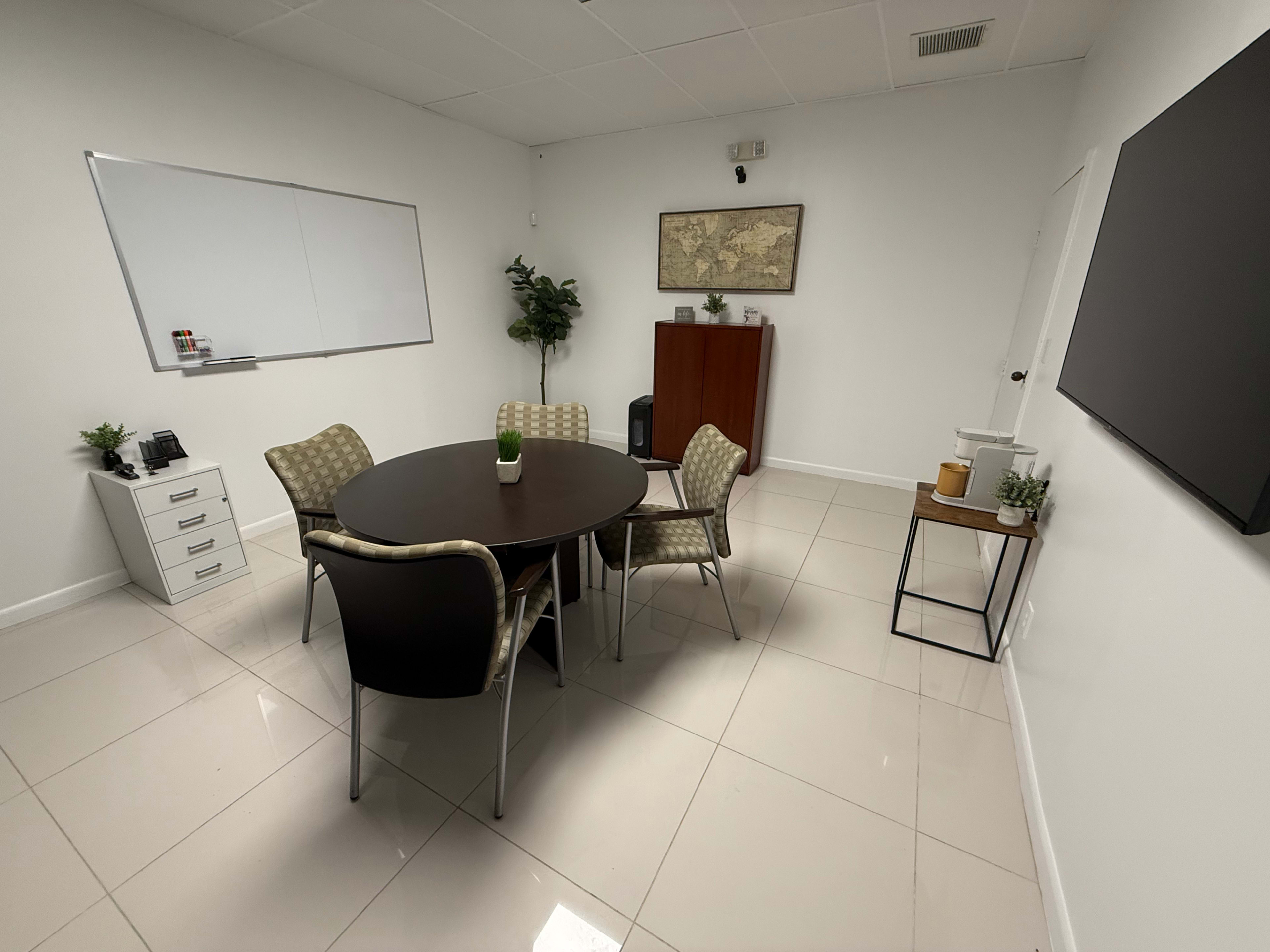 The image shows a modern conference room with a round table surrounded by four chairs, a whiteboard on one wall, a map on another, and a few decorative plants.