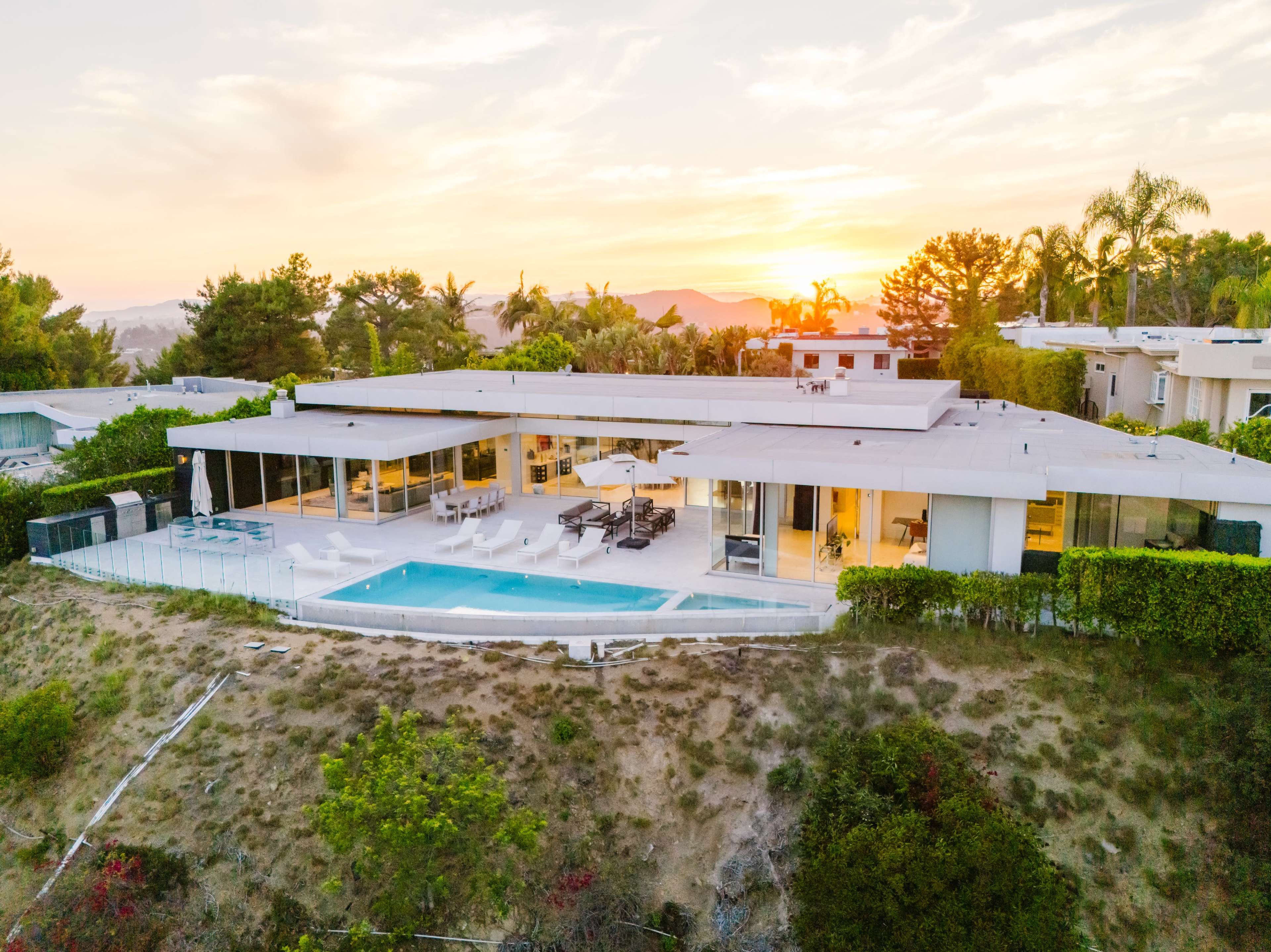A modern single-story house with a swimming pool is situated on a hillside, accompanied by palm trees and a sunset background.