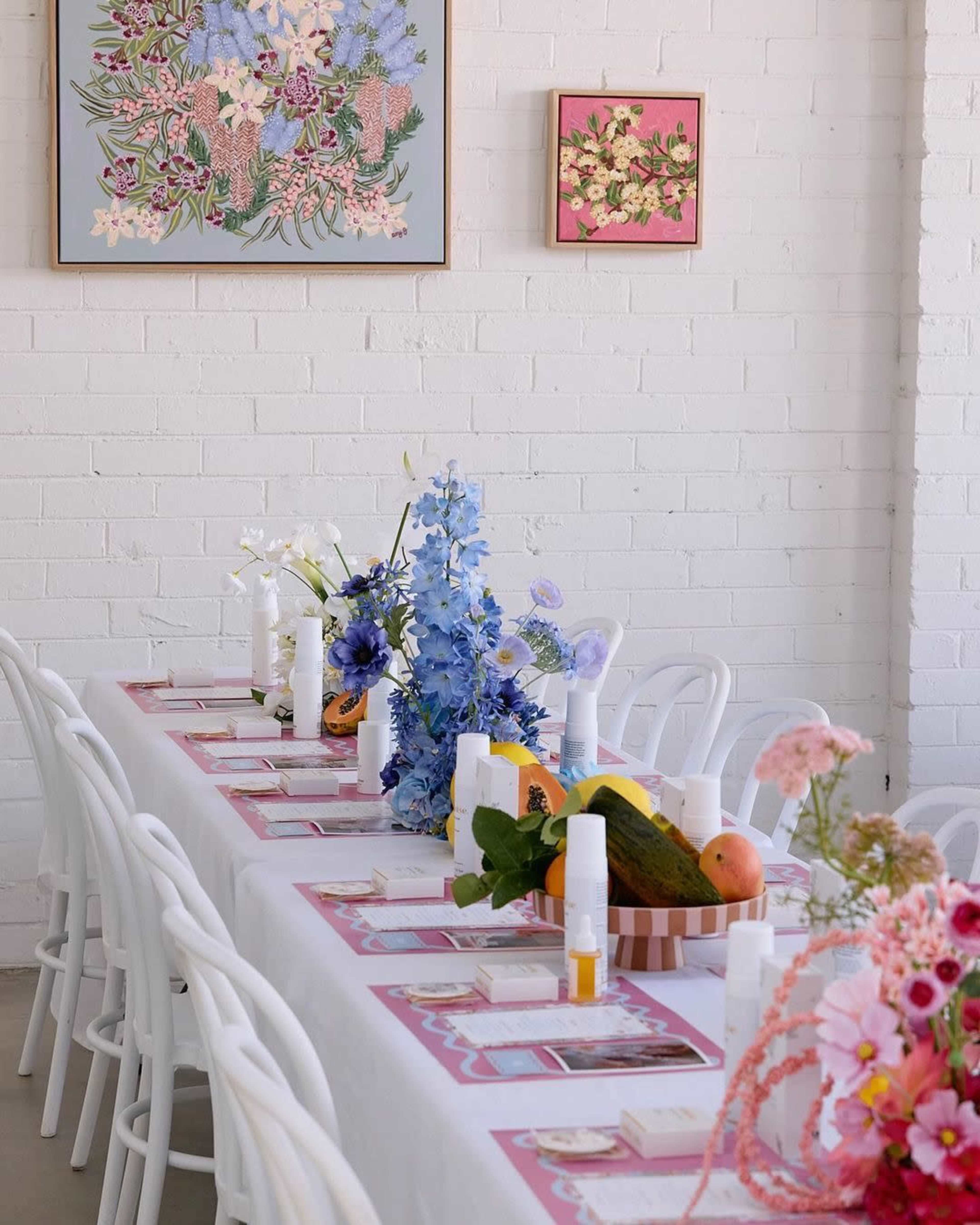 A long dining table is set with floral centerpieces and skincare products, against a backdrop of white brick walls and floral art.