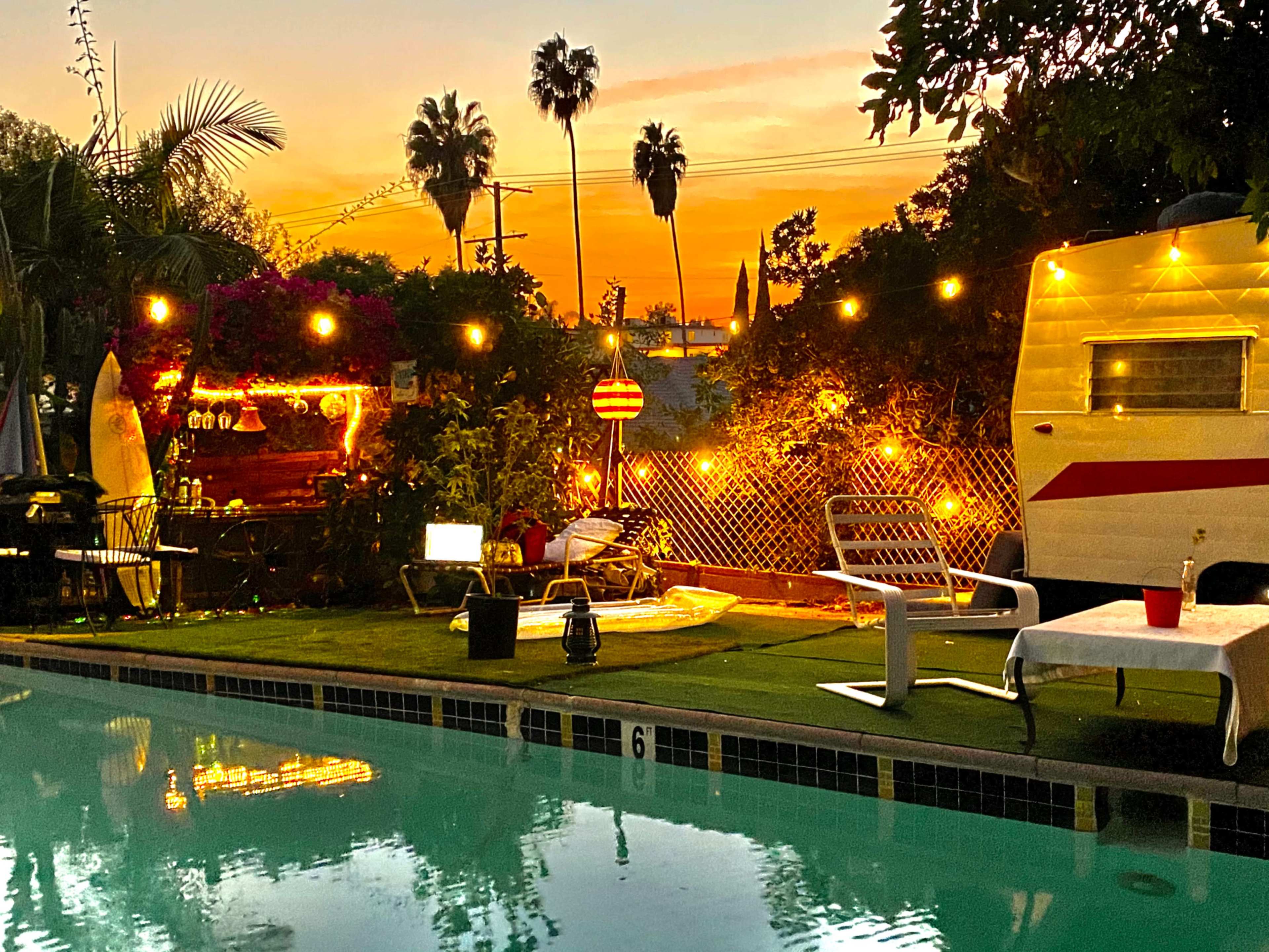 A swimming pool is illuminated by string lights, with a vintage camper, lounge chairs, and tropical plants in the background during sunset.