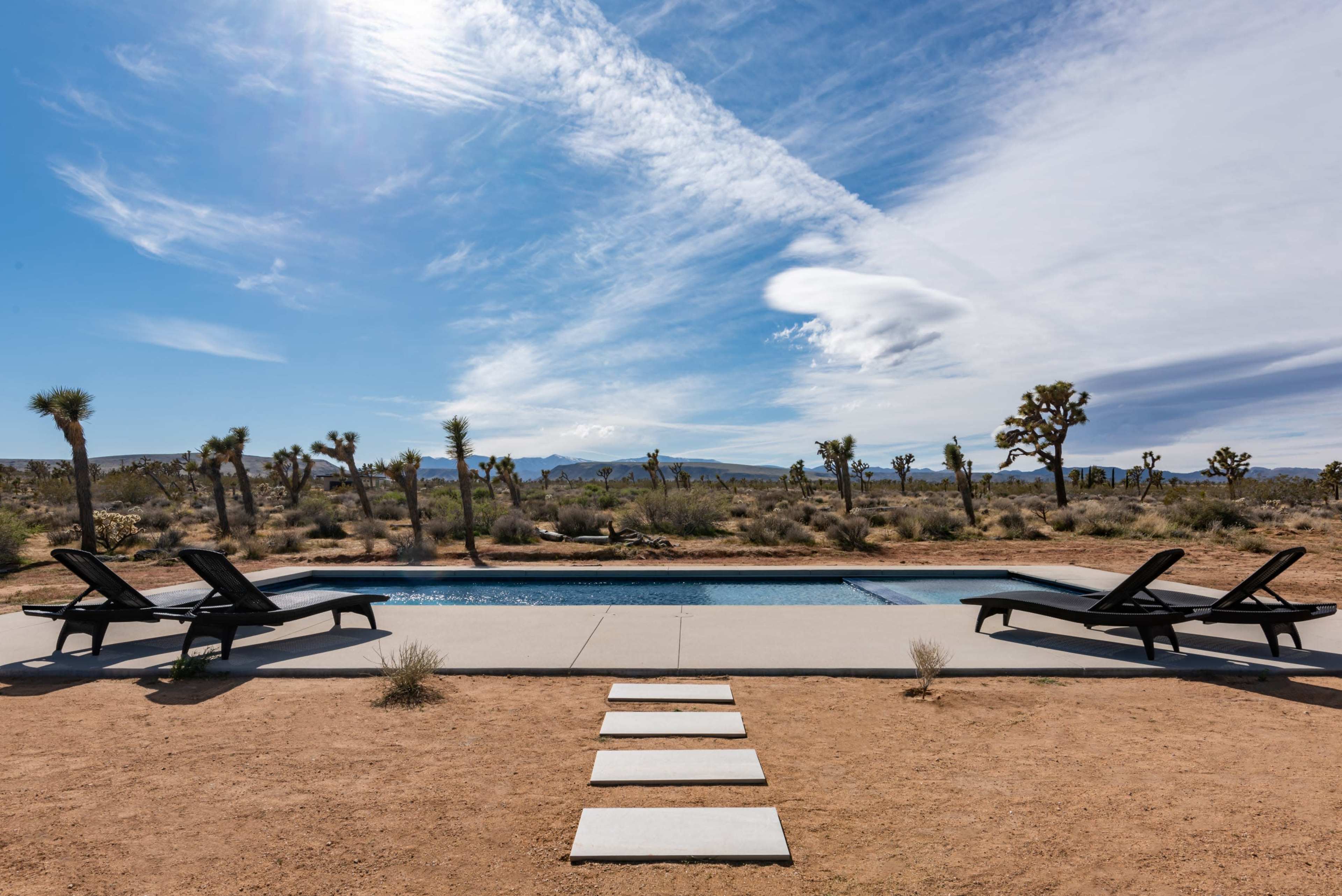 A rectangular swimming pool is surrounded by desert landscaping and lounge chairs, with Joshua trees dotting the background under a blue sky.