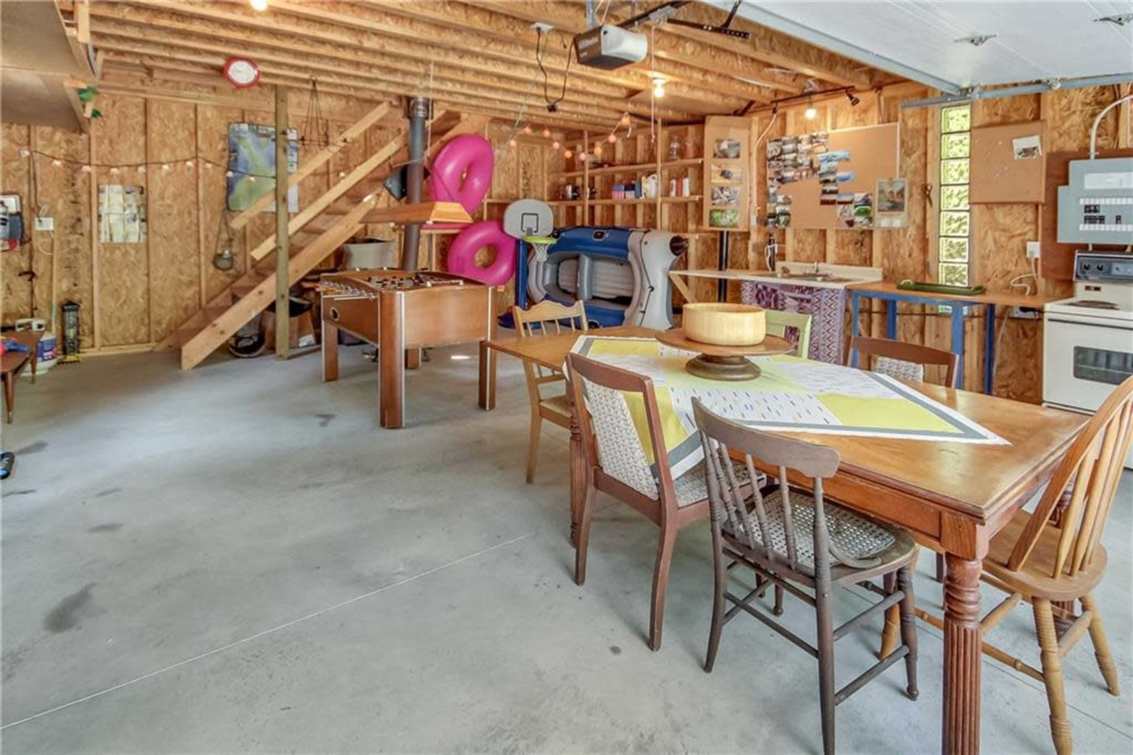 The image shows a spacious garage with wooden walls, featuring a dining table, chairs, a foosball table, and various recreational equipment.