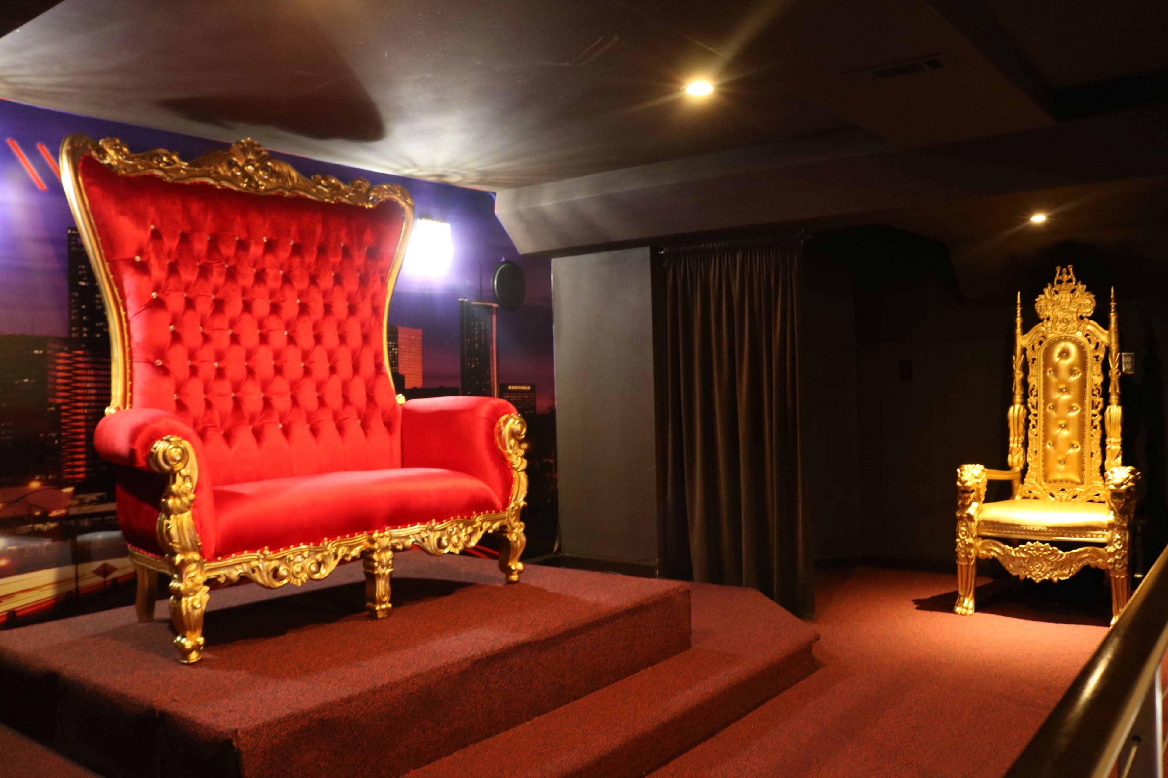 The image shows a lavish red velvet throne with gold accents alongside an ornate golden chair, both positioned on a raised platform in an indoor setting.