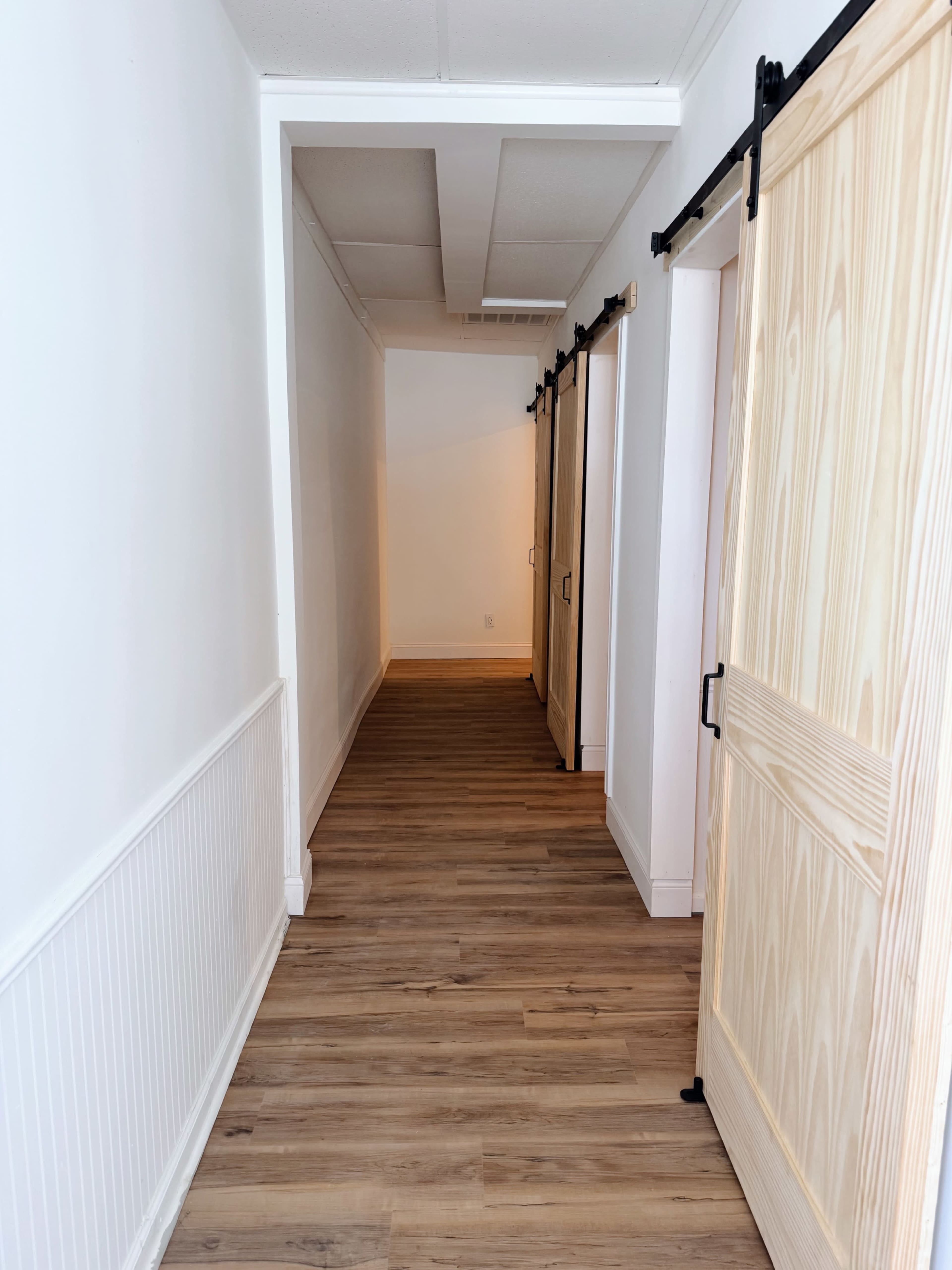 A narrow hallway features multiple wooden sliding doors along its sides, with light-colored flooring and white walls.