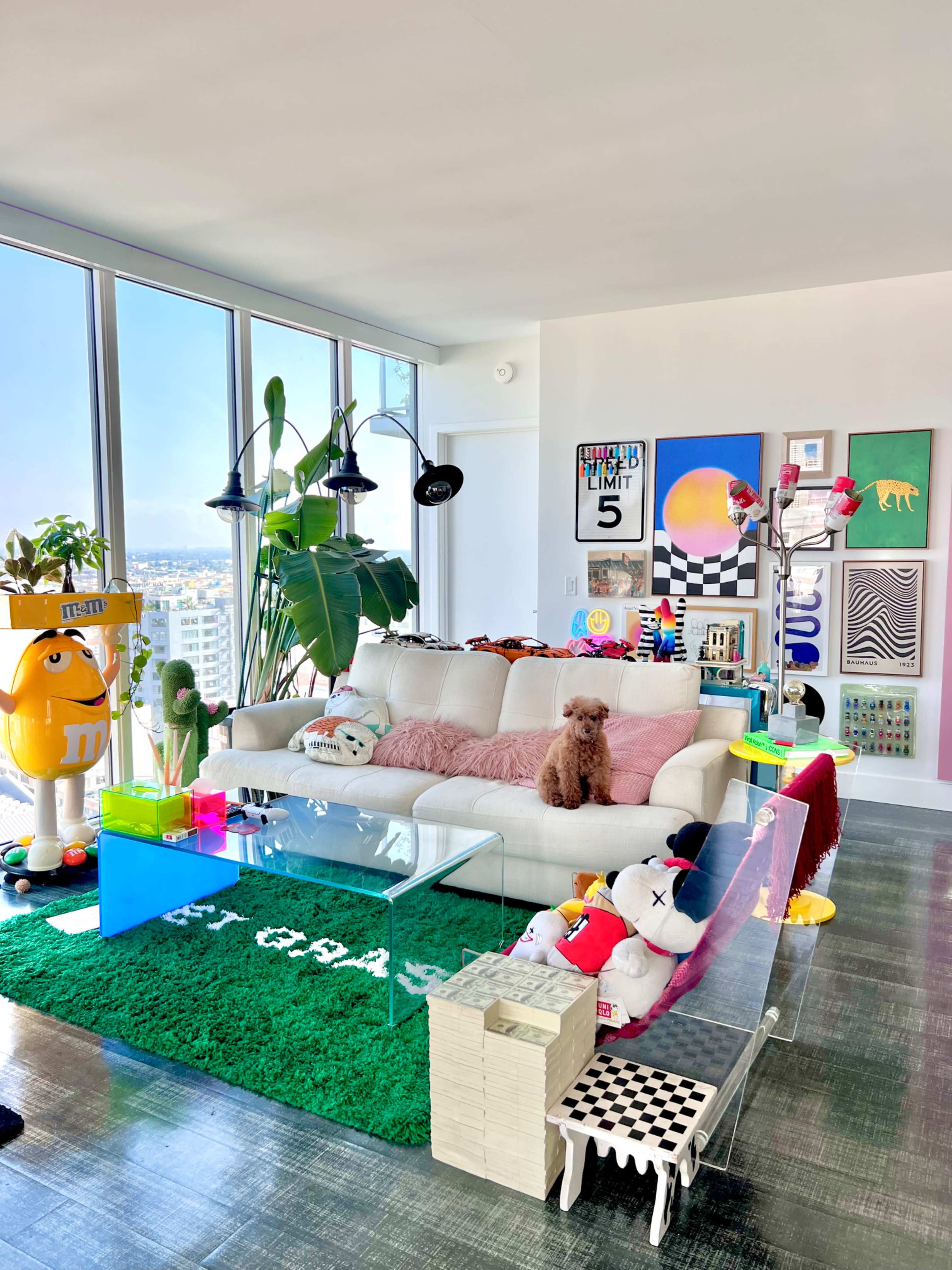 A modern living room features a white sofa adorned with colorful cushions, a clear coffee table, and various playful decorations, all set against large windows showcasing a city view.