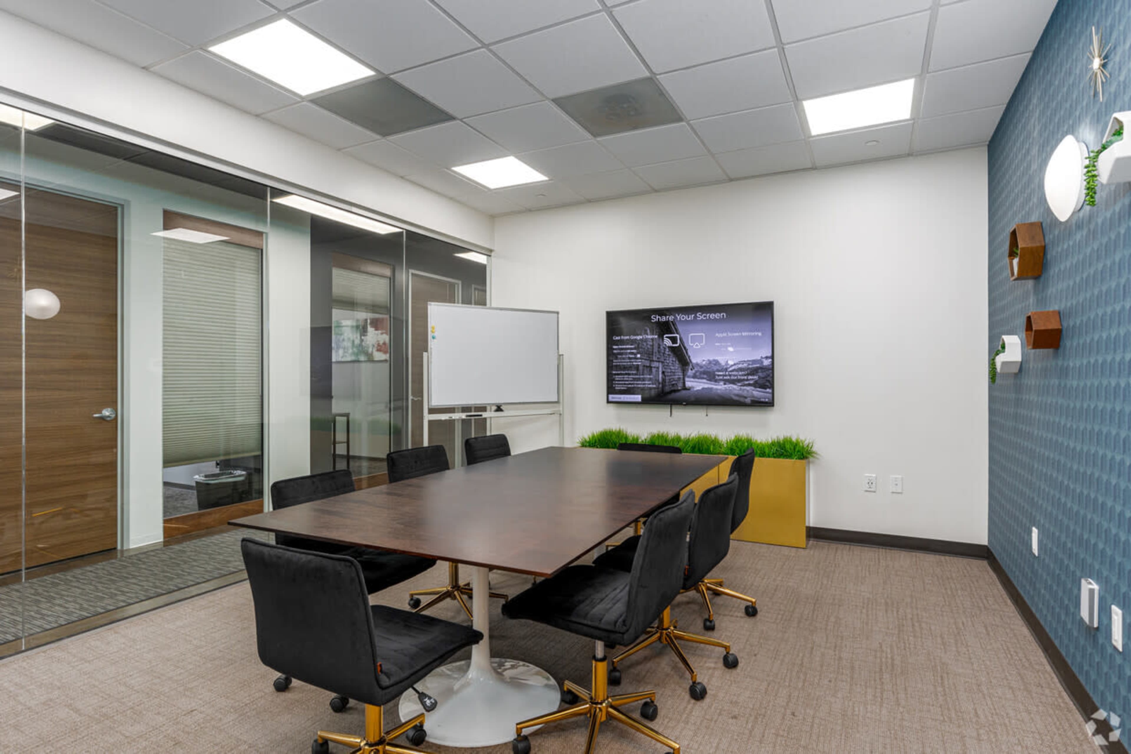 A modern conference room features a large wooden table surrounded by black chairs, with a wall-mounted screen displaying content and glass partitioning.