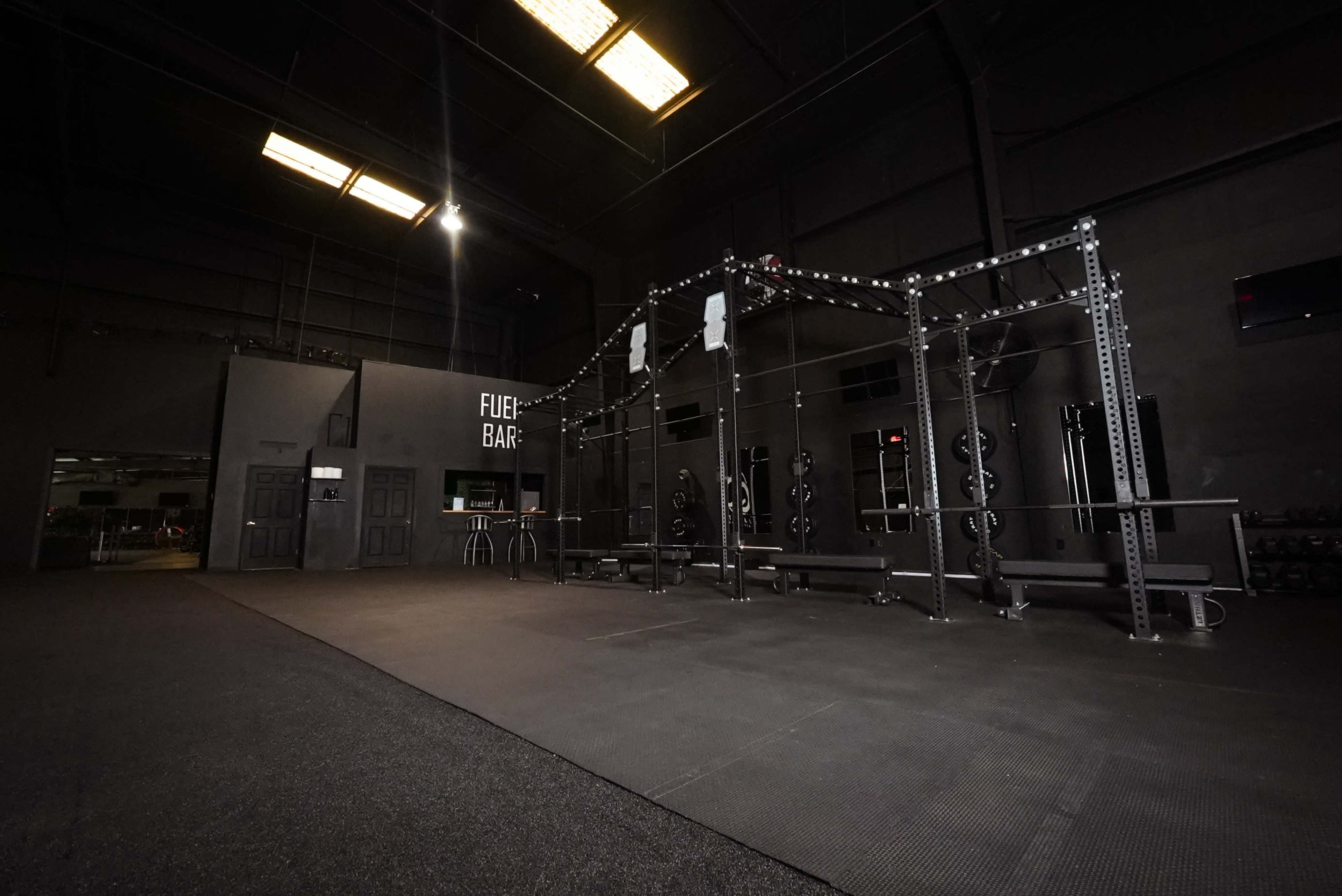 The image shows a spacious, dimly lit gym area featuring a metal weight rack, benches, and workout equipment against a dark wall.