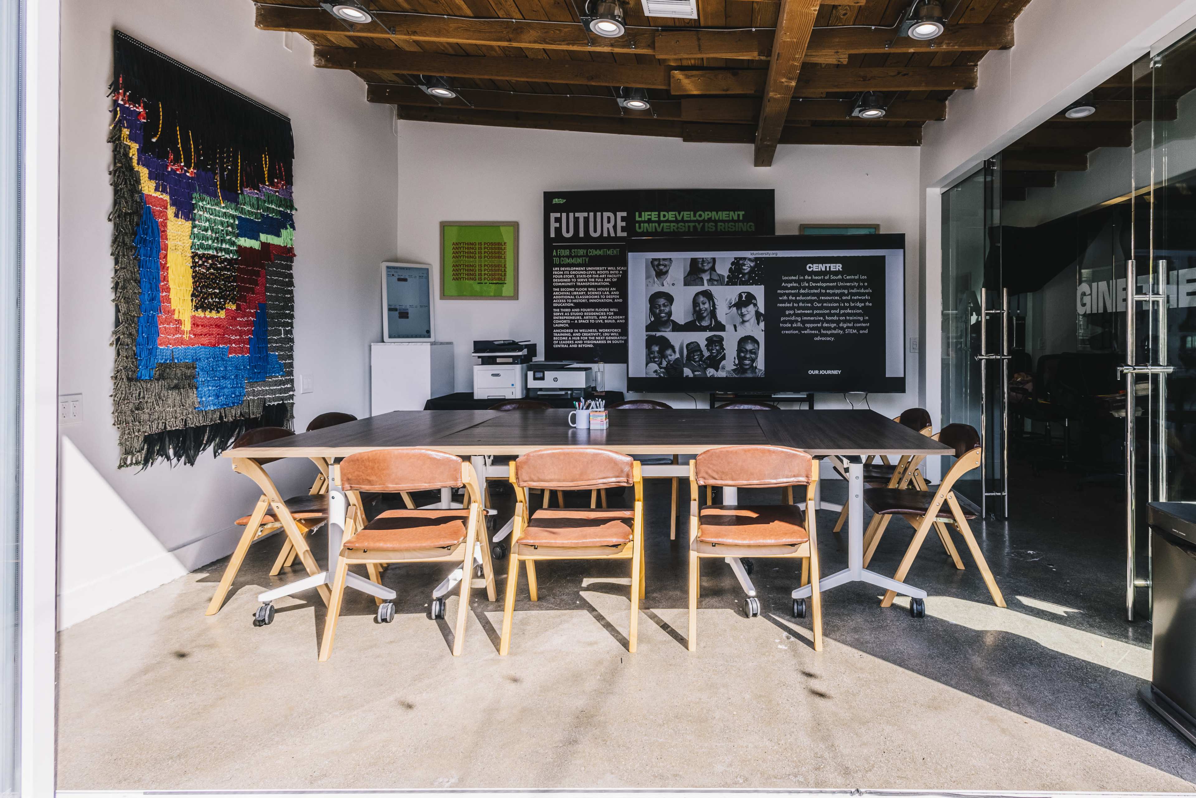 The image shows a modern conference room with a large table surrounded by wooden chairs, featuring a large screen displaying information on a wall and colorful artwork in the background.