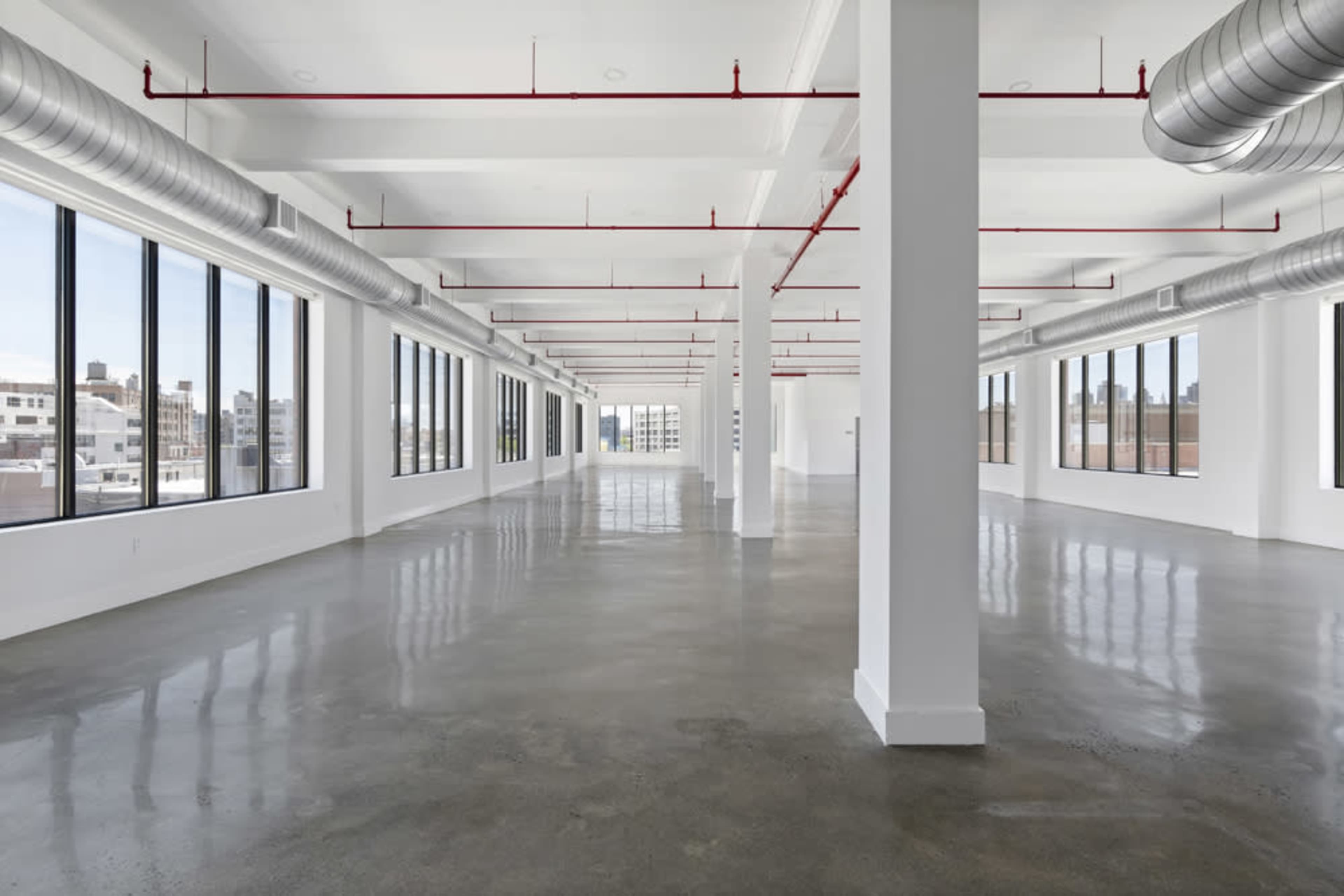 A spacious, unfinished commercial interior with concrete flooring and large windows, featuring exposed red piping along the ceiling.