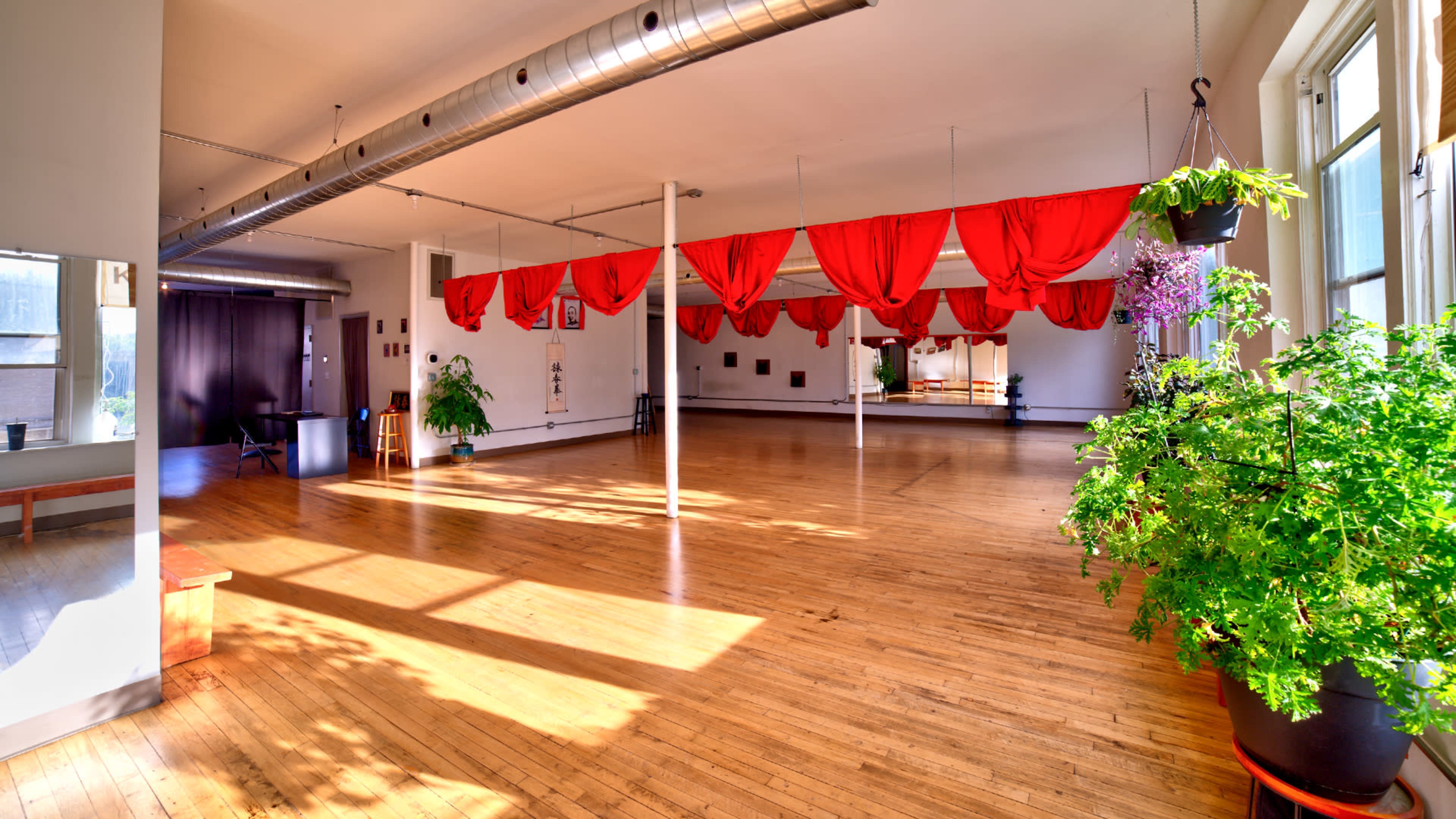 The image shows a spacious studio with wooden flooring, large windows, and red fabric drapes hanging from the ceiling, complemented by potted plants.