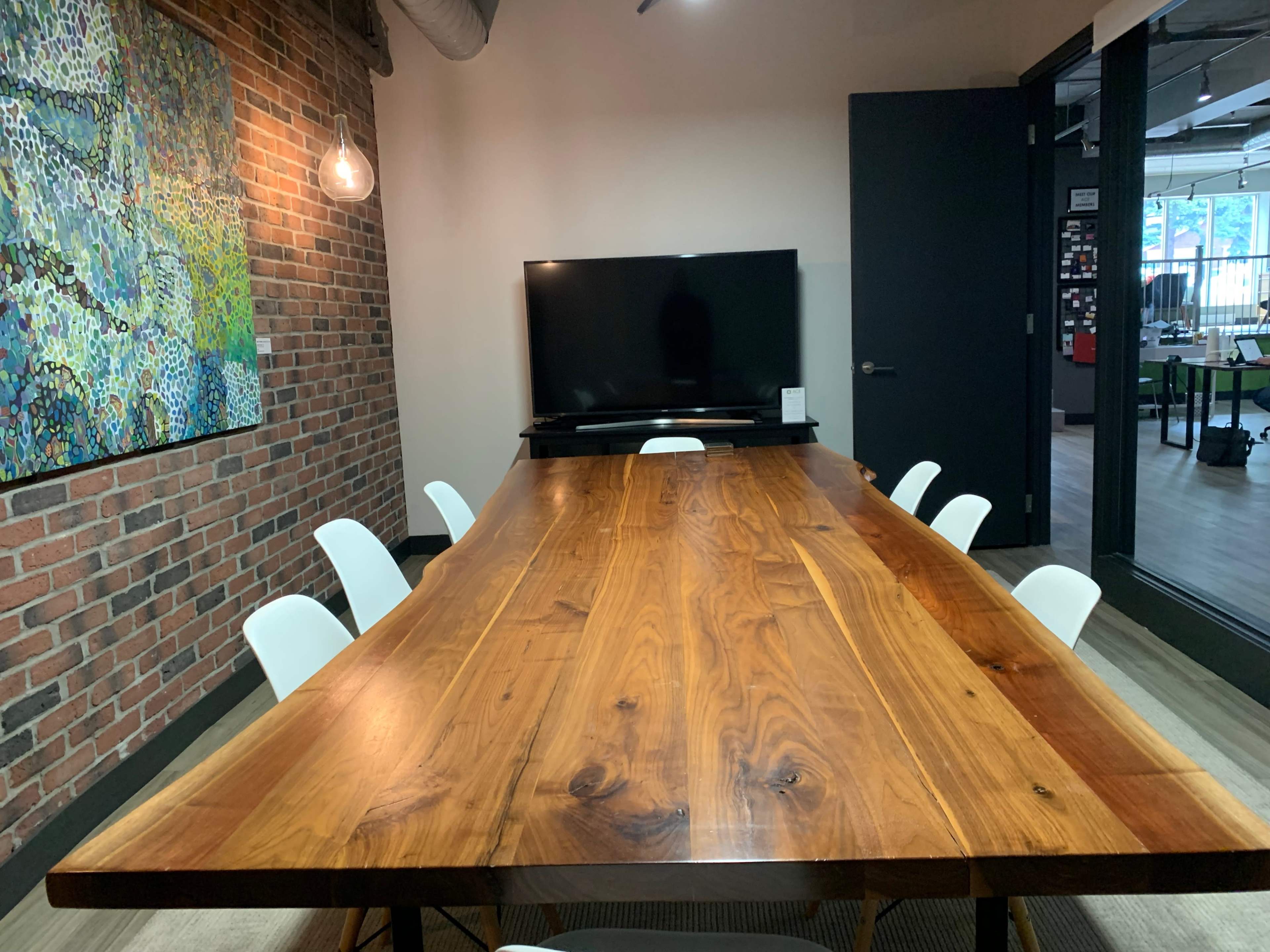 A large wooden table is positioned in a meeting room with a brick wall, surrounded by white chairs, and a television mounted on the opposing wall.