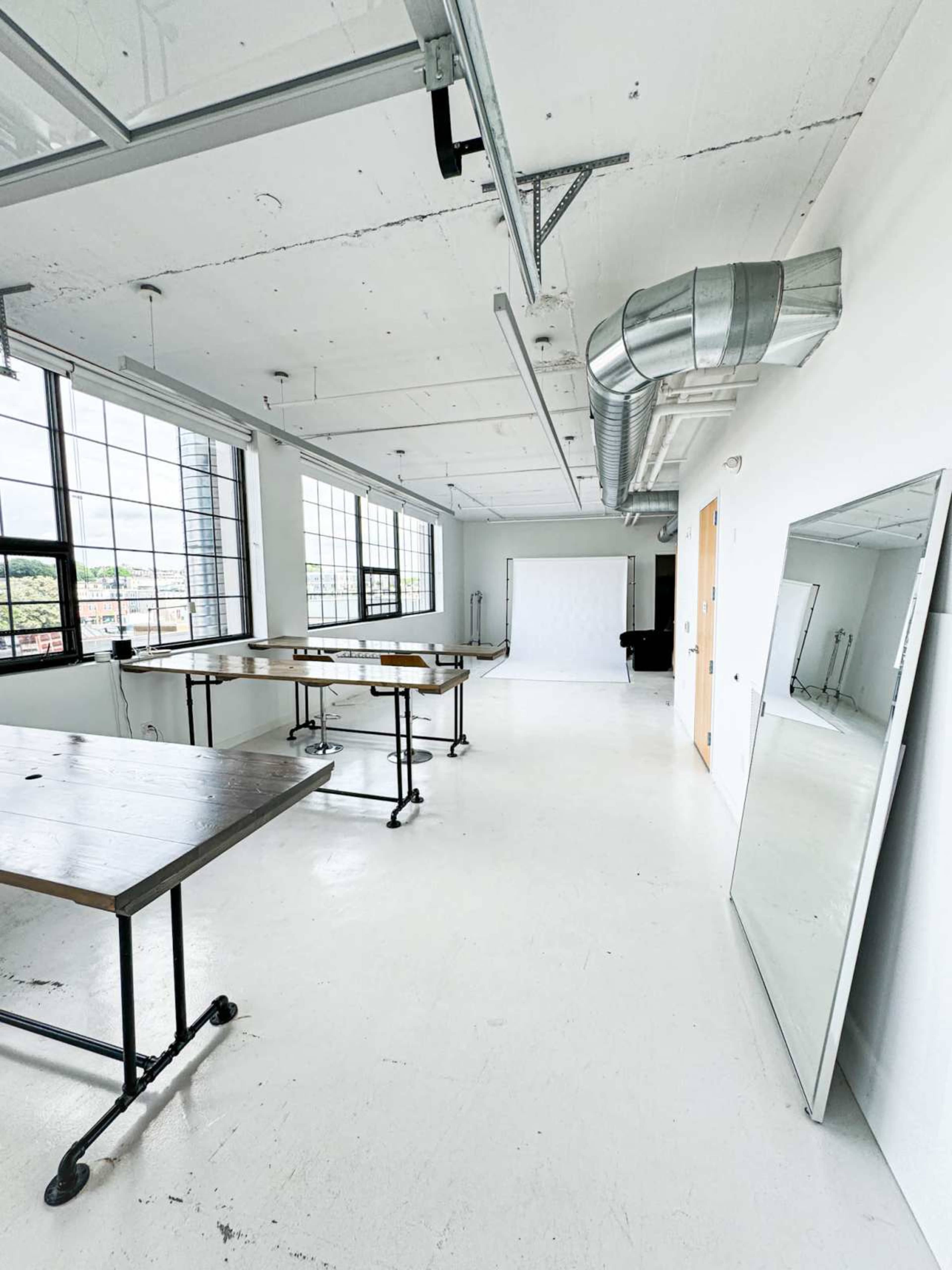 The image shows an empty studio space with large windows, wooden tables on wheels, and a white backdrop.