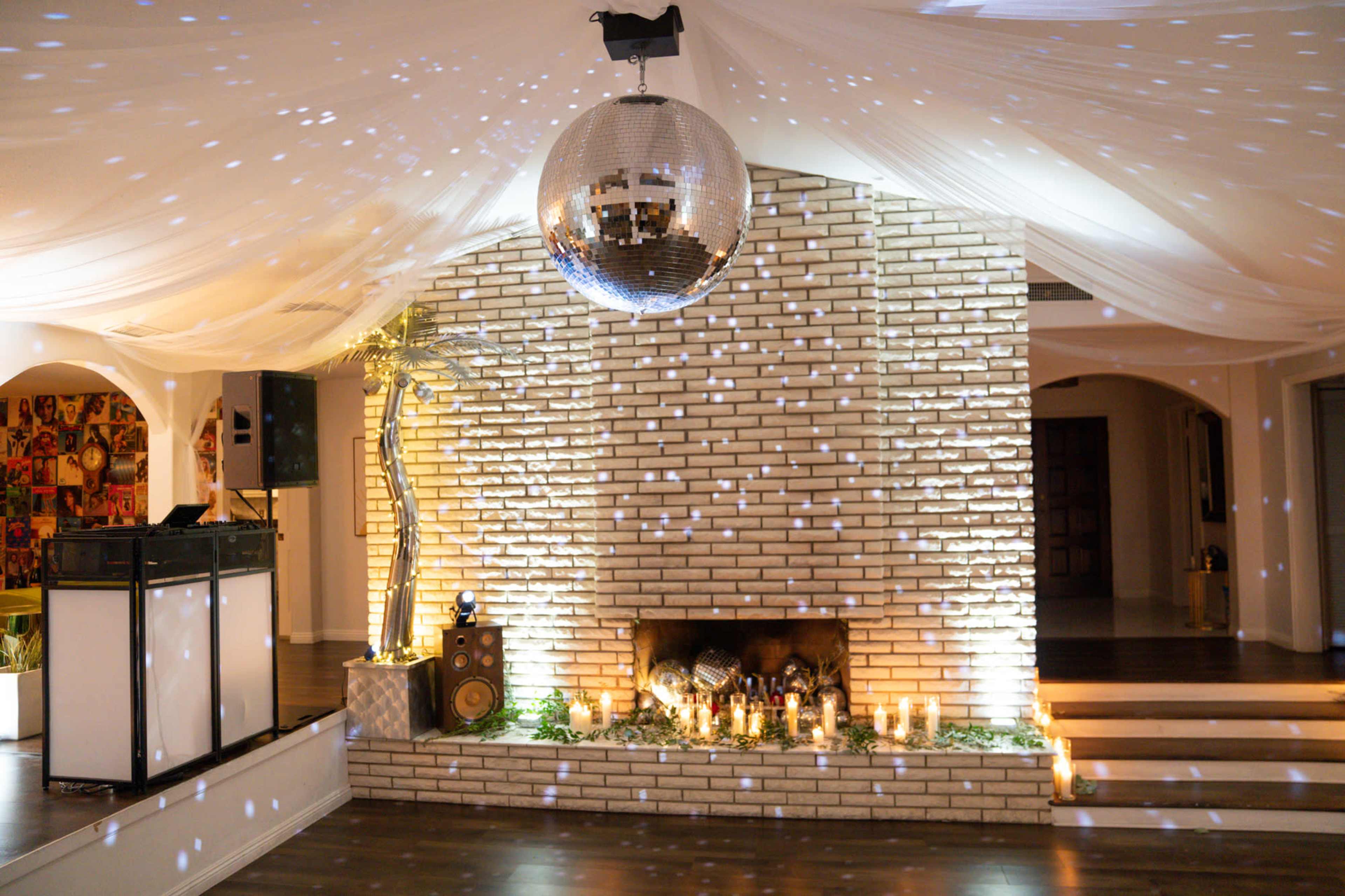 The image shows a decorated indoor event space featuring a large disco ball suspended above a brick fireplace adorned with candles and greenery.