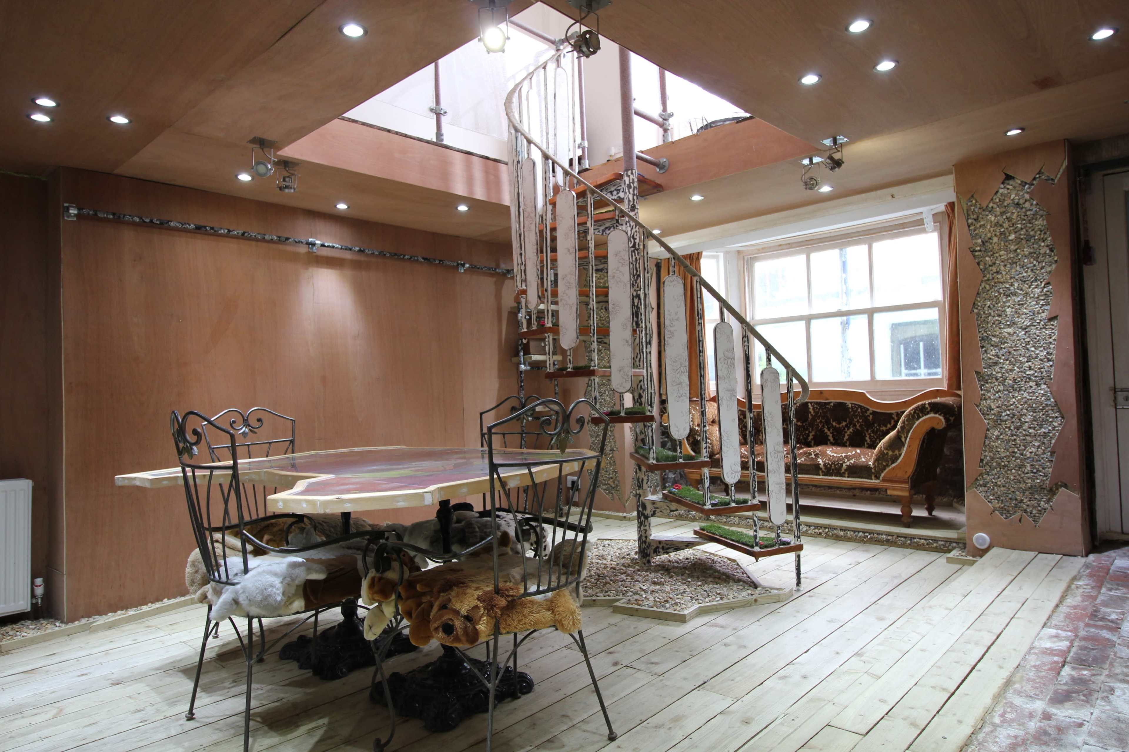 An interior space featuring a spiral staircase, a round dining table with chairs, and a furnished sitting area with a couch, all designed with wooden elements and a mix of materials.