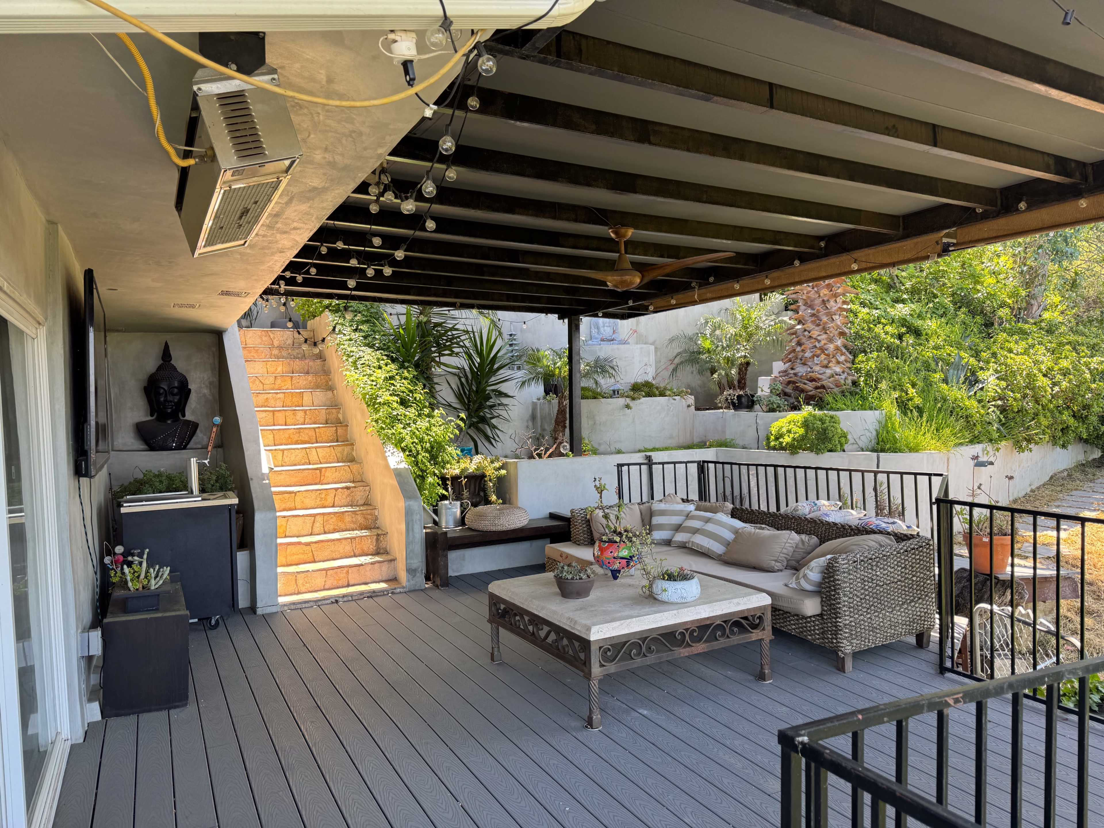 A covered patio features a seating area with a coffee table, surrounded by greenery and a staircase leading to an upper level.