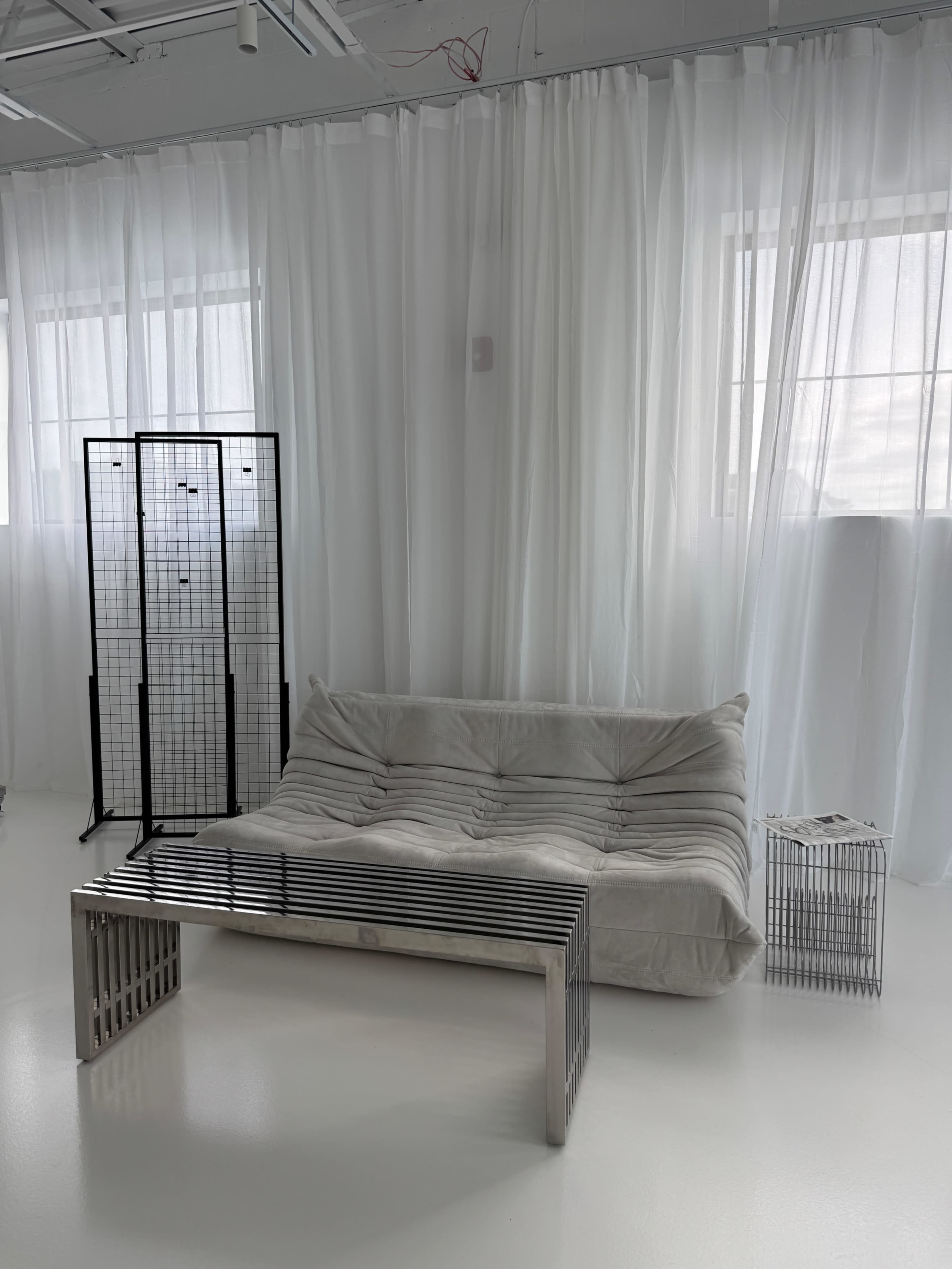 The image shows a minimalist living space featuring a light-colored sofa, a metal coffee table, and a room divider, all set against sheer white curtains and large windows.