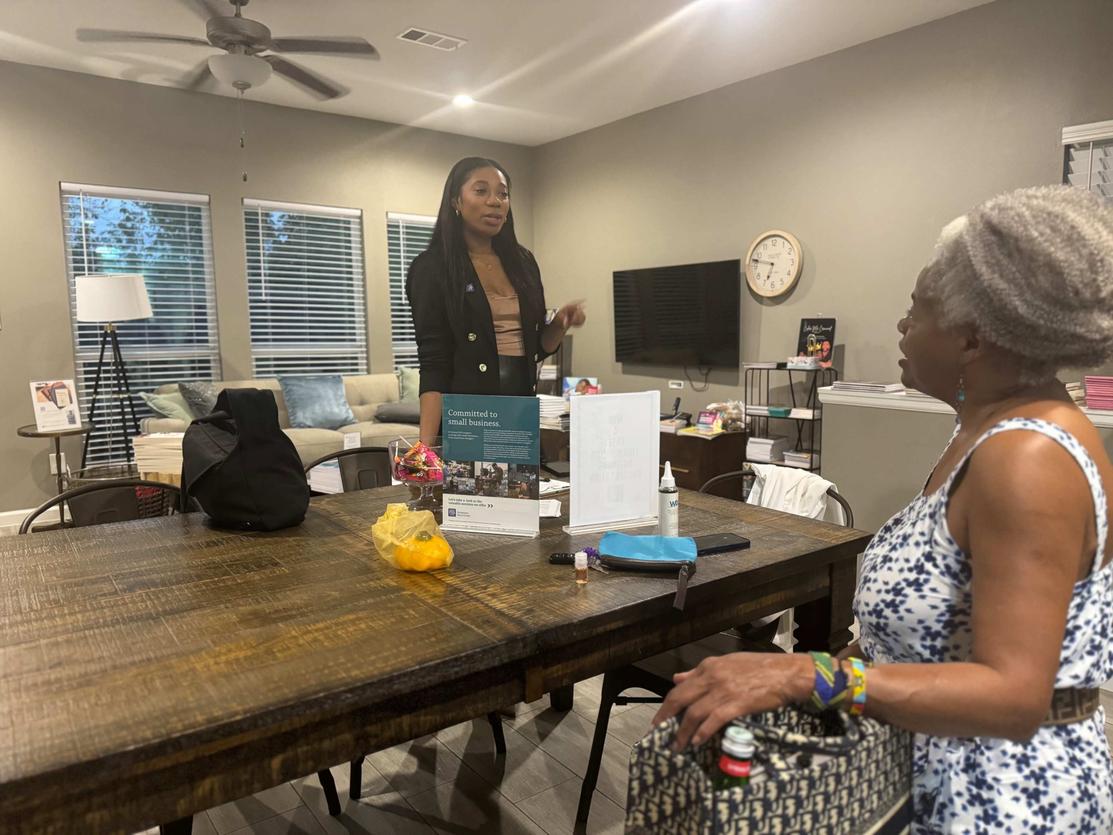 A woman in a black blazer speaks to another woman seated at a wooden table in a well-lit living space.
