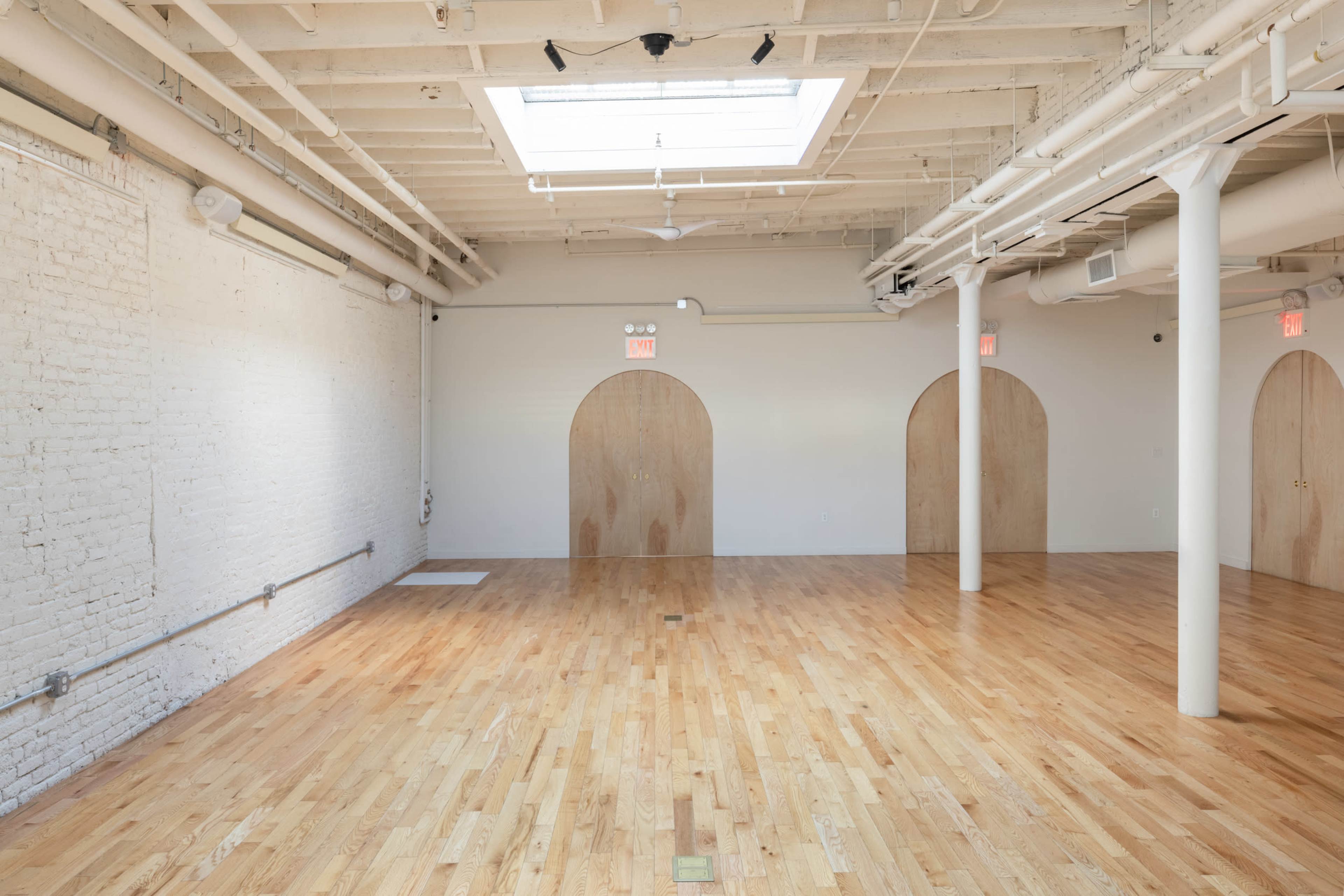 An empty, well-lit room with wooden floors, white brick walls, and three arched wooden doors.