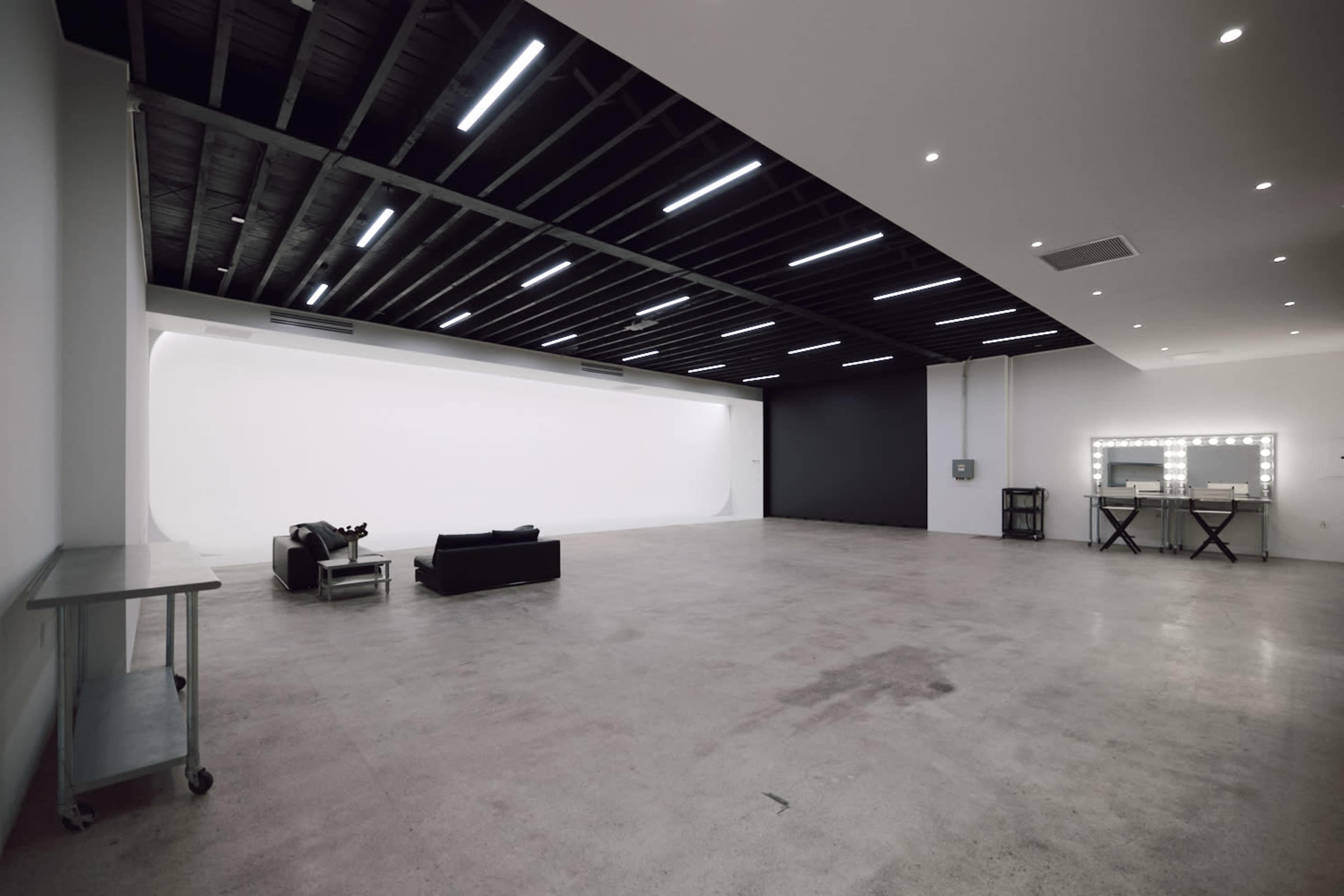 The image shows a spacious, empty studio with a minimalist design, featuring a black wall, a white backdrop, a couple of chairs, and a vanity mirror with lights.