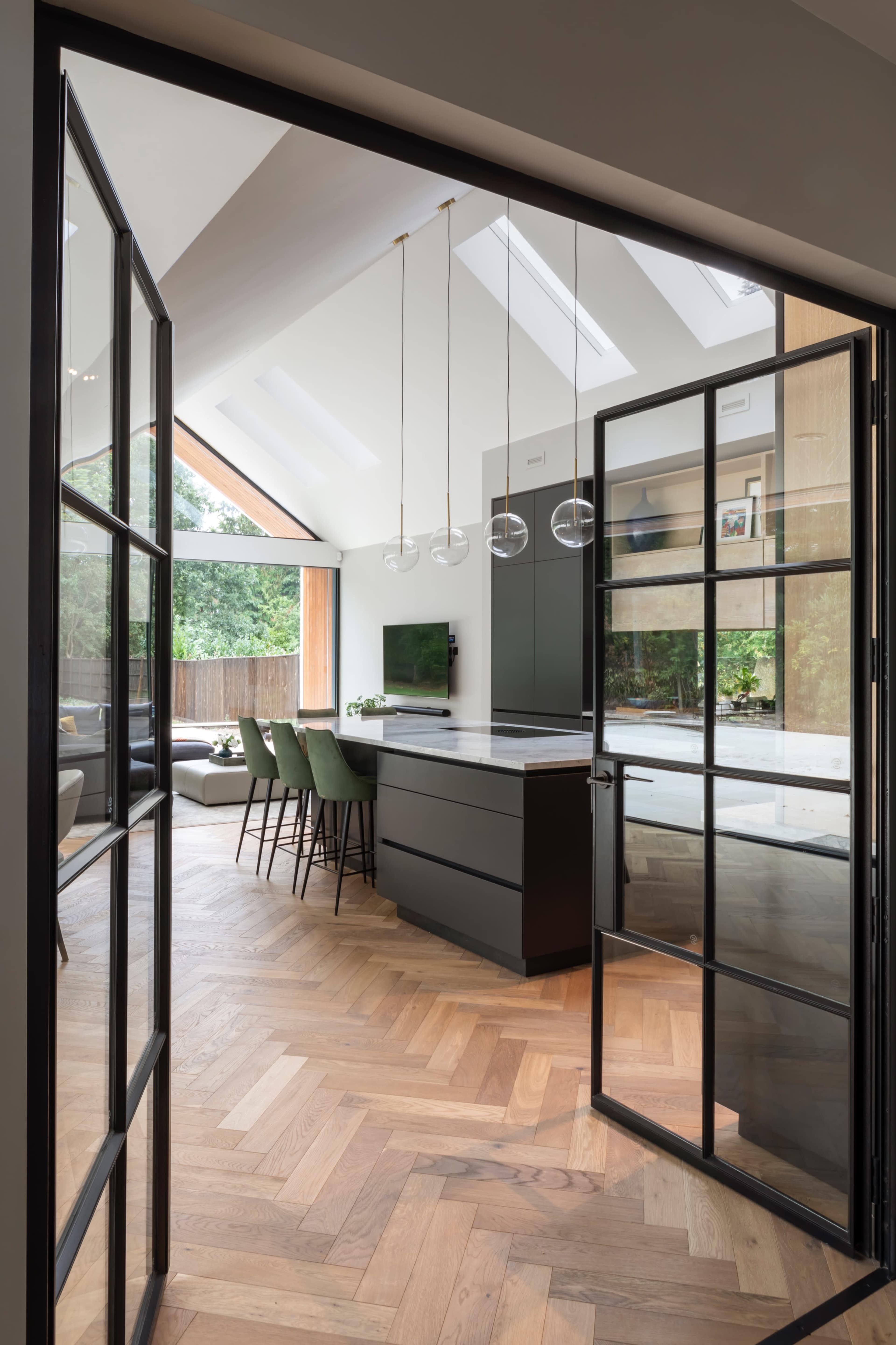 A modern kitchen with an open layout features a large island, bar stools, and skylights, framed by black glass doors.