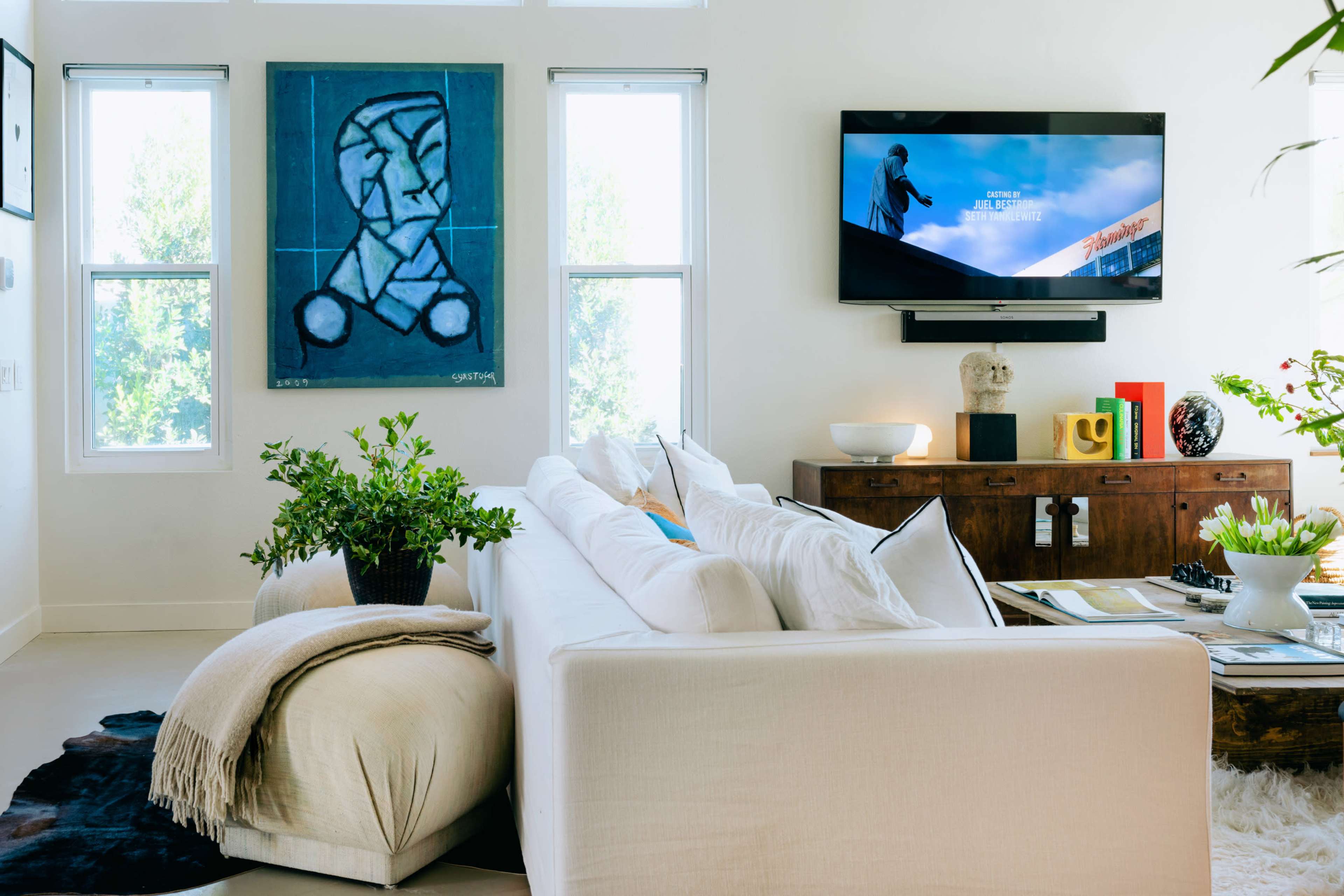 A modern living room features a white sofa, a large abstract painting on the wall, a television mounted above a wooden console, and a variety of decorative plants.