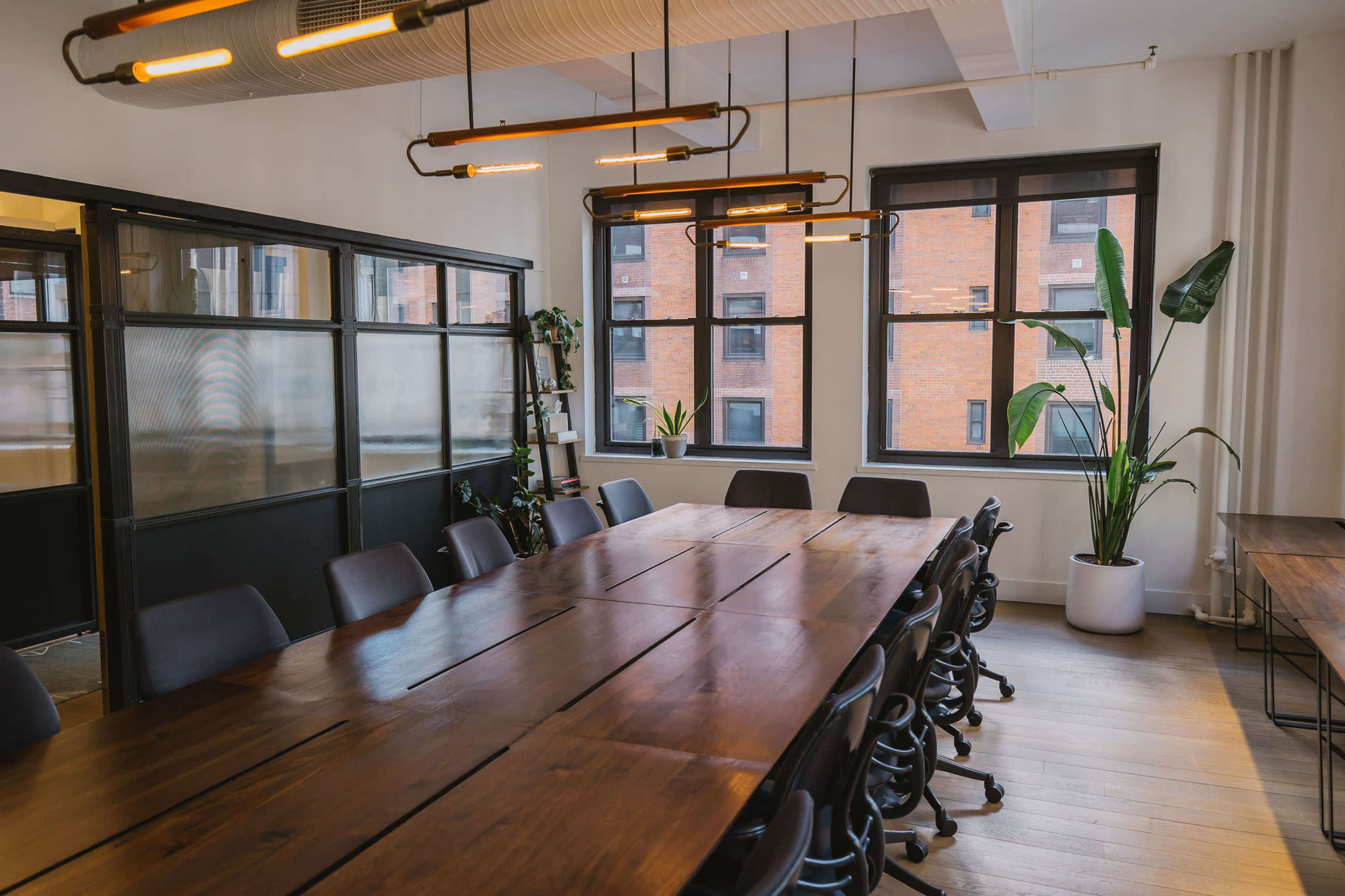 A long wooden conference table with chairs is placed in a well-lit meeting room featuring large windows and decorative plants.