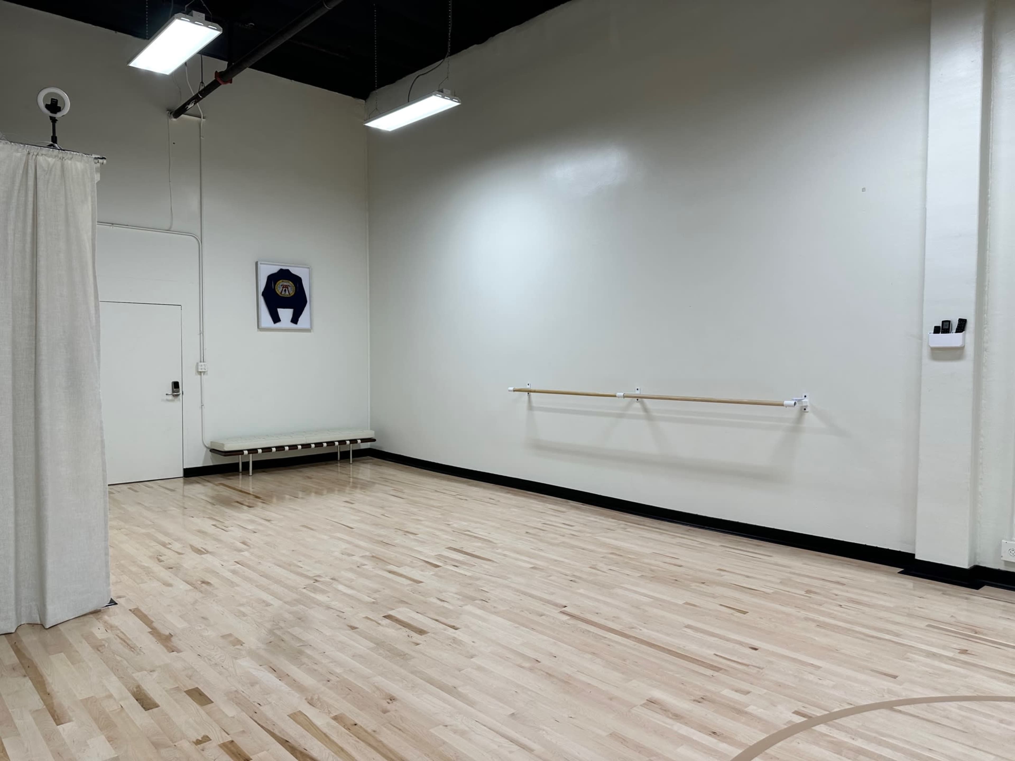 The image shows a spacious dance studio with wooden flooring, a wall-mounted ballet barre, and minimal furnishings.