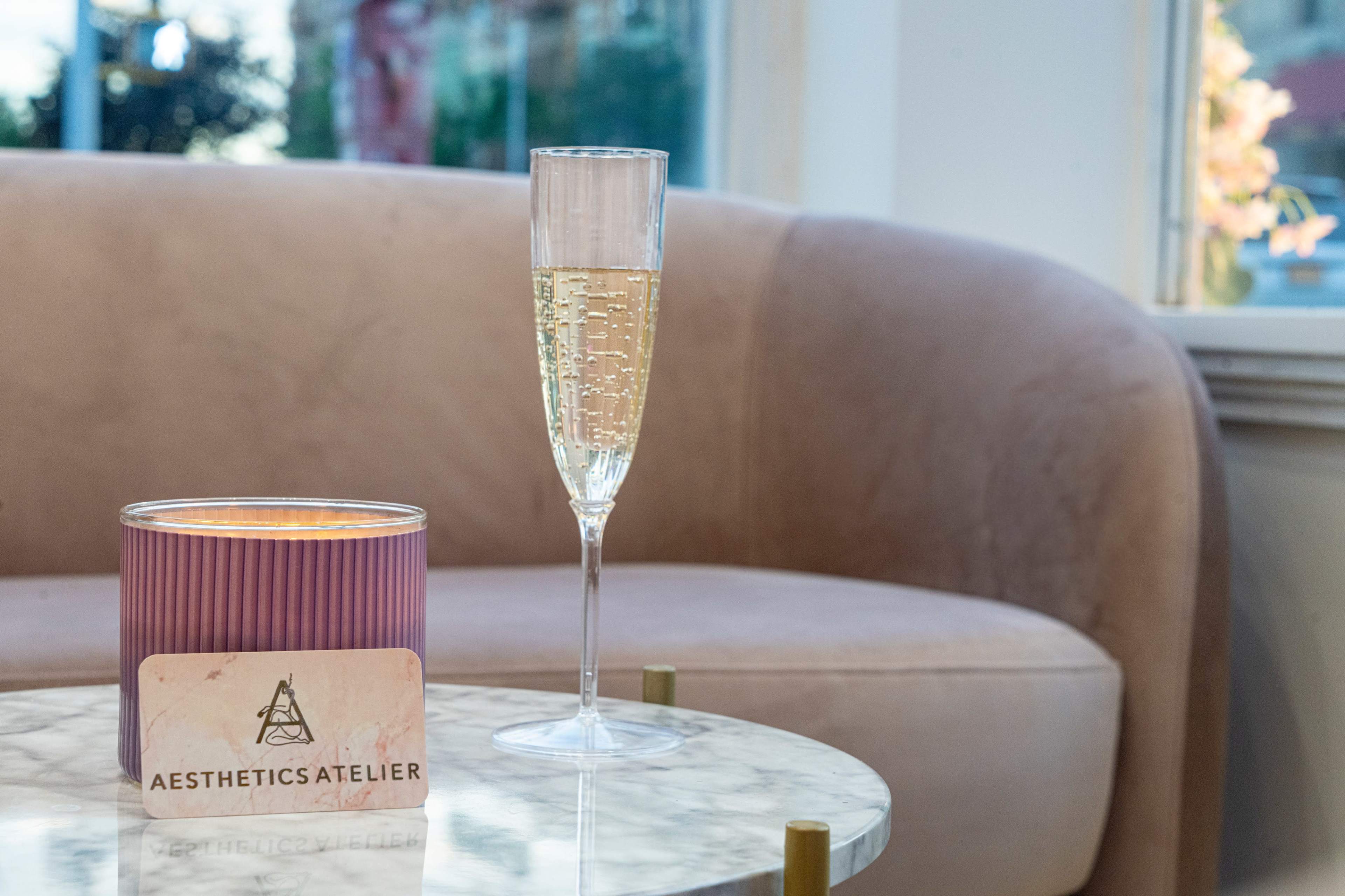 A clear champagne flute sits beside a lit candle on a marble table, with a plush beige sofa in the background.