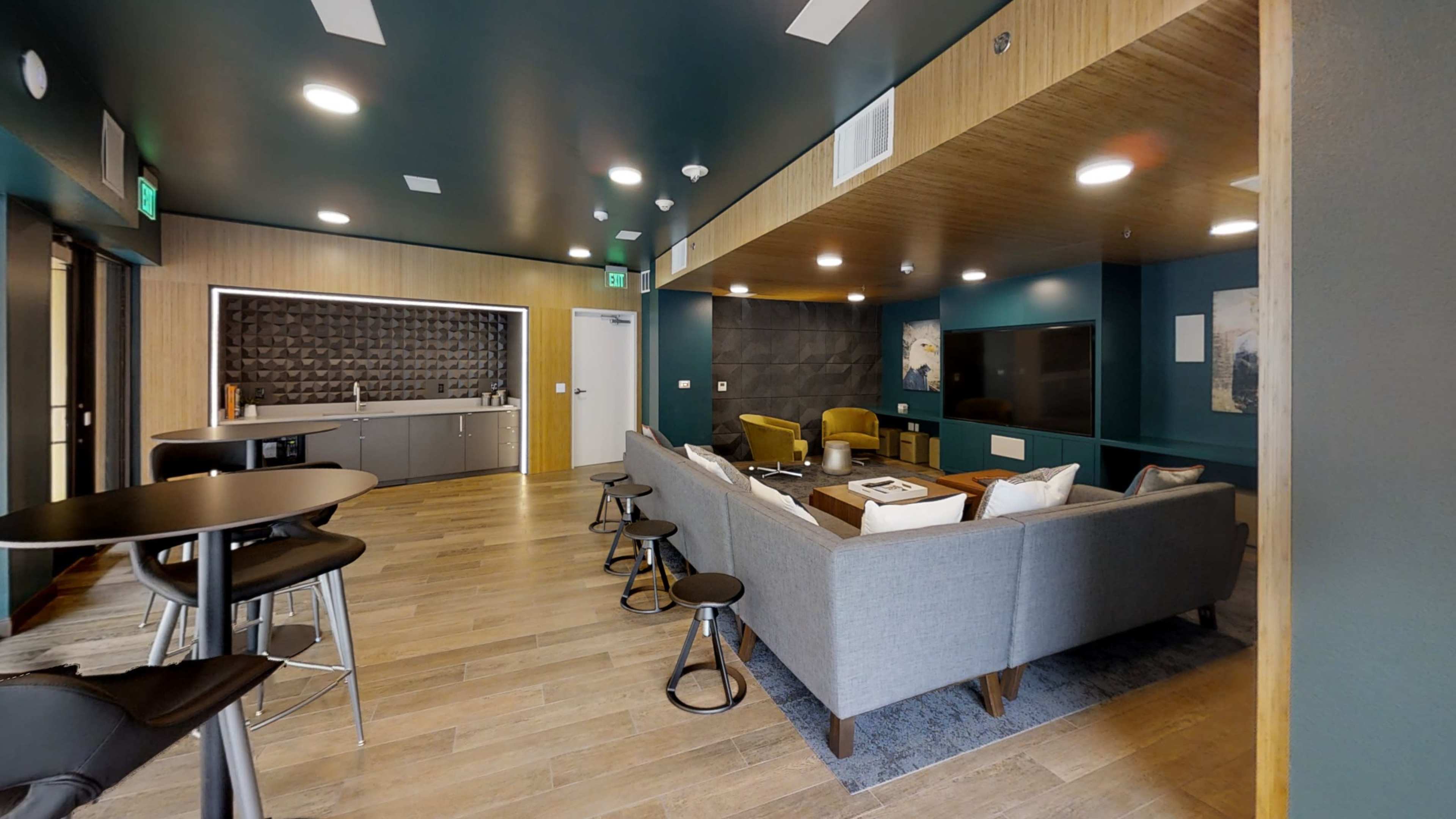 A modern lounge area featuring a seating arrangement with a gray sofa, two yellow chairs, a kitchenette, and a dark accent wall with a TV.