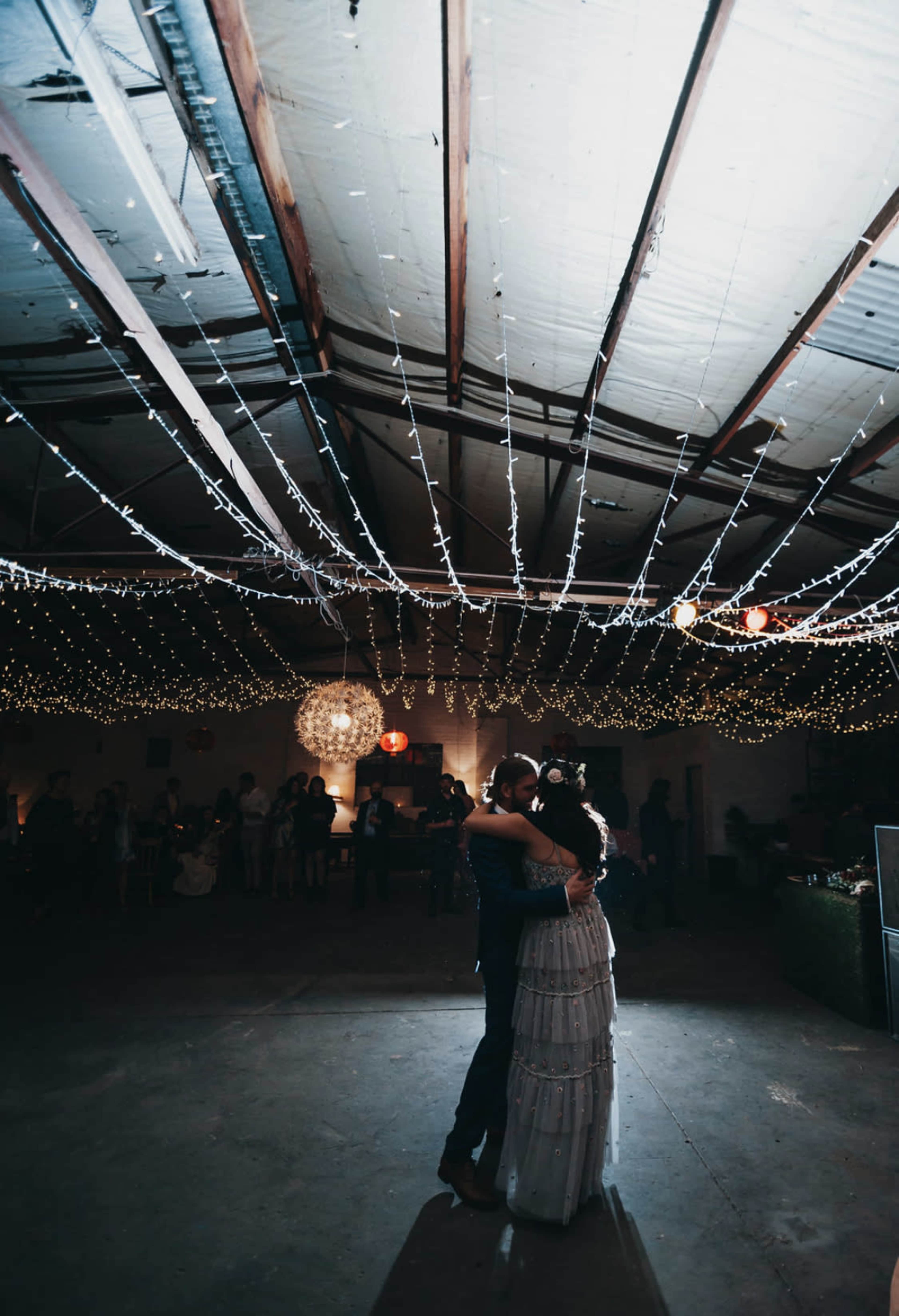 A couple dances together under a canopy of string lights in a spacious venue with guests in the background.