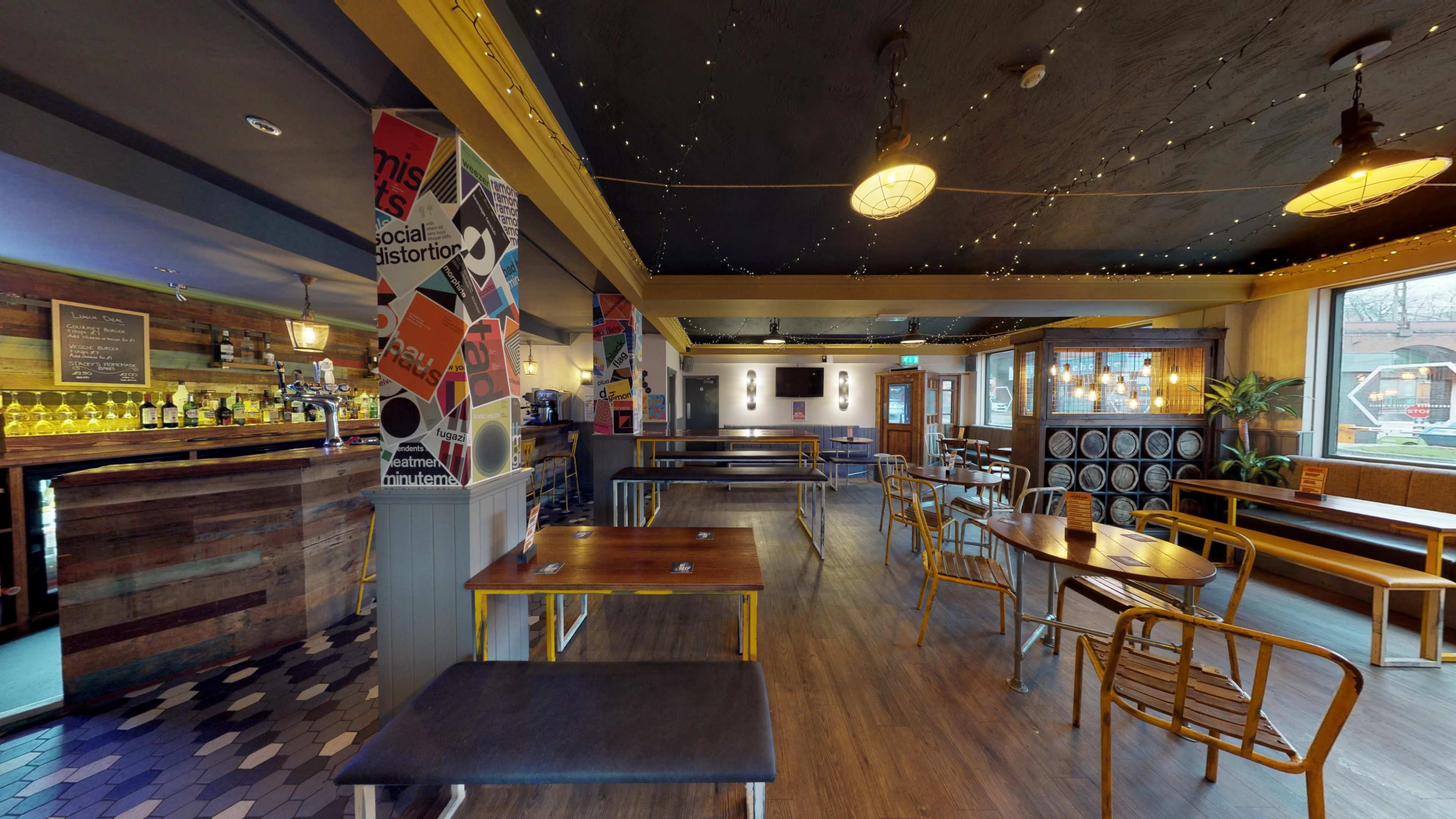 The image shows a spacious pub interior with wooden tables, benches, a bar stocked with bottles, and decorative posters on the walls.