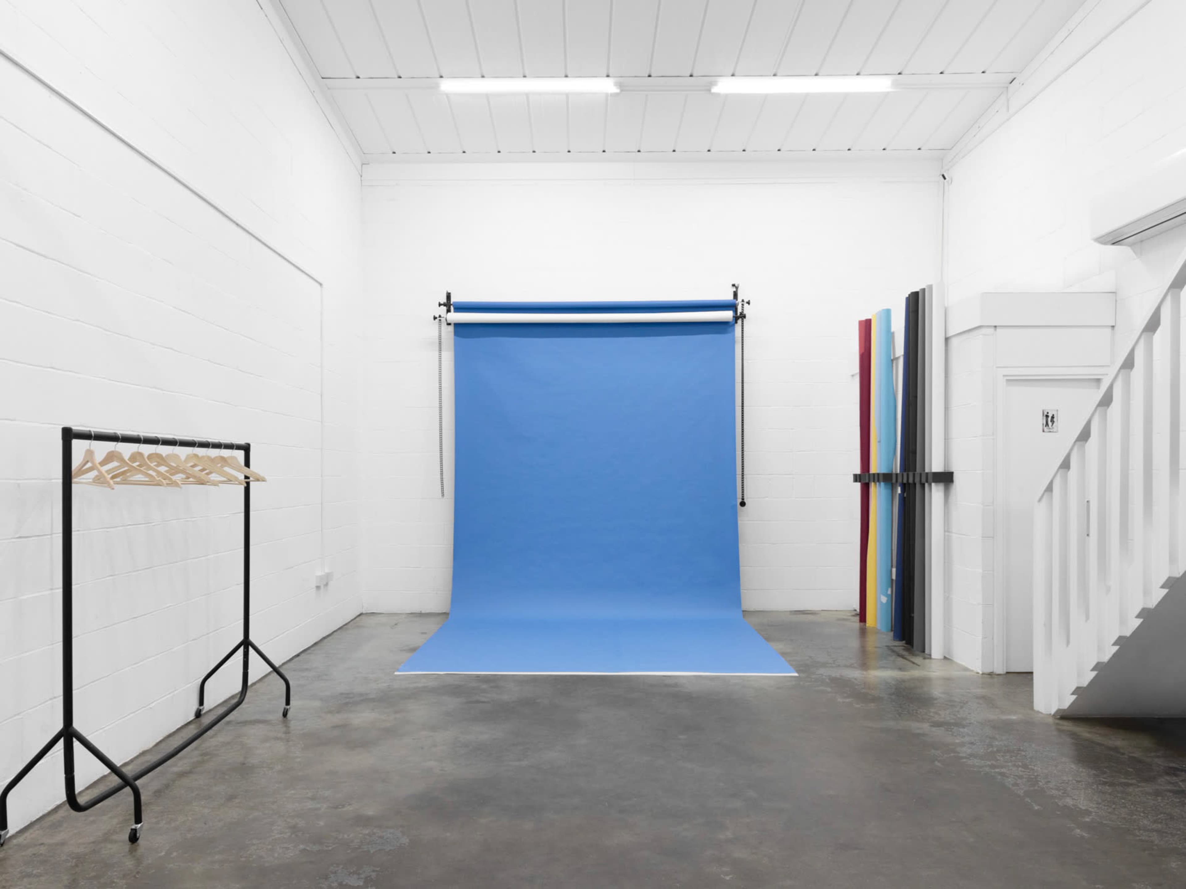 A photography studio features a blue backdrop in the center, accompanied by a clothing rack with hangers and colorful backdrops leaning against the wall.