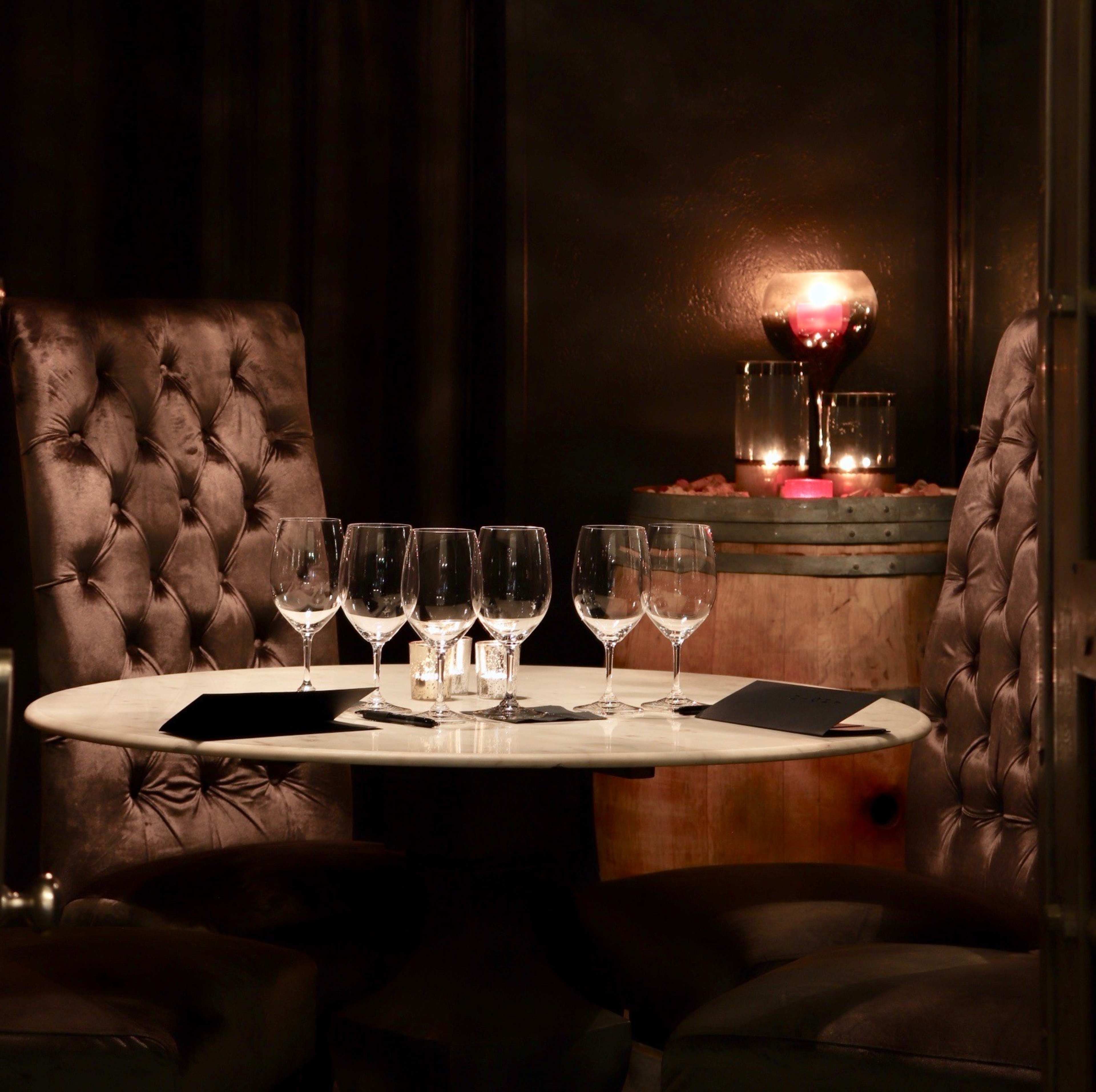 A marble table with six glasses and black menus is set between two tufted chairs, with a candle-lit centerpiece in the background.