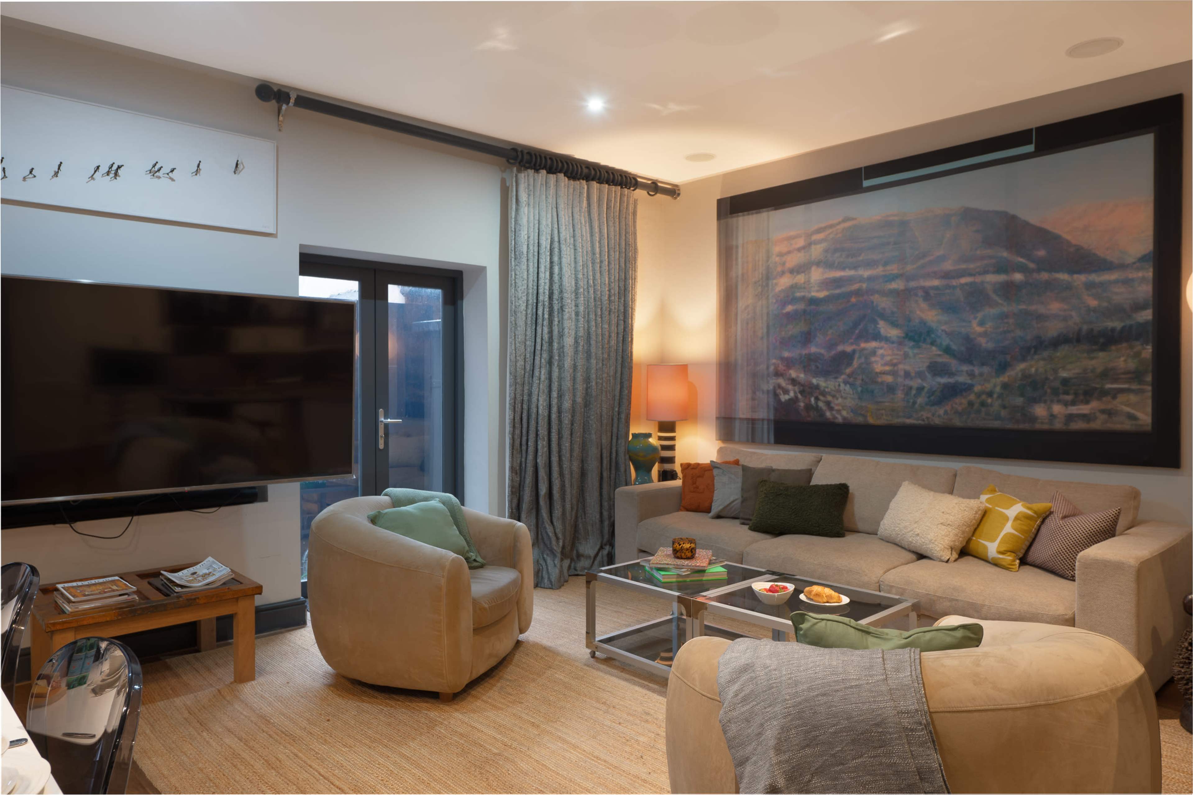 The image depicts a modern living room featuring a sectional sofa, two armchairs, a large television, and a wall-mounted artwork.