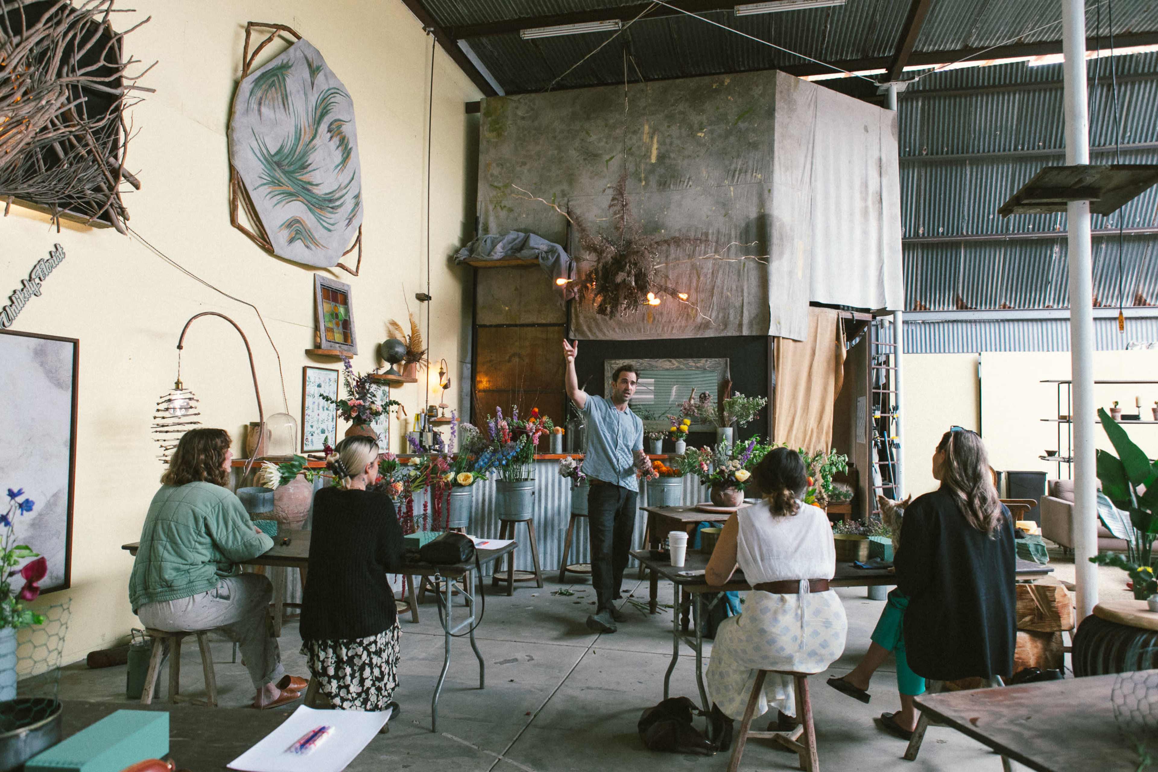 A group of people is engaged in a floral design workshop inside a spacious, decorated industrial venue.