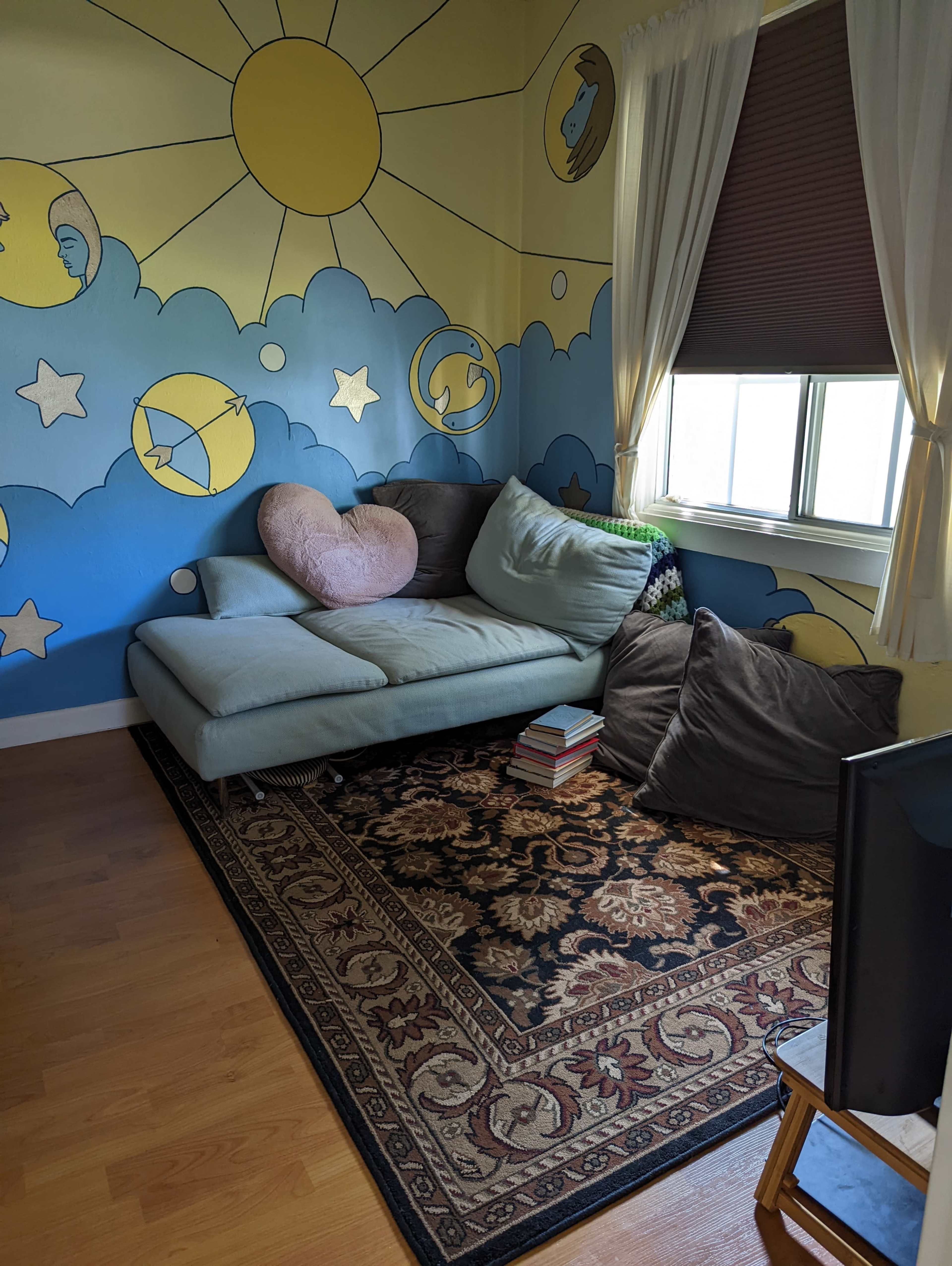 The image shows a cozy corner of a room with a light blue sofa, large pillows, a decorative rug, and a colorful mural featuring sun and cloud designs on the walls.