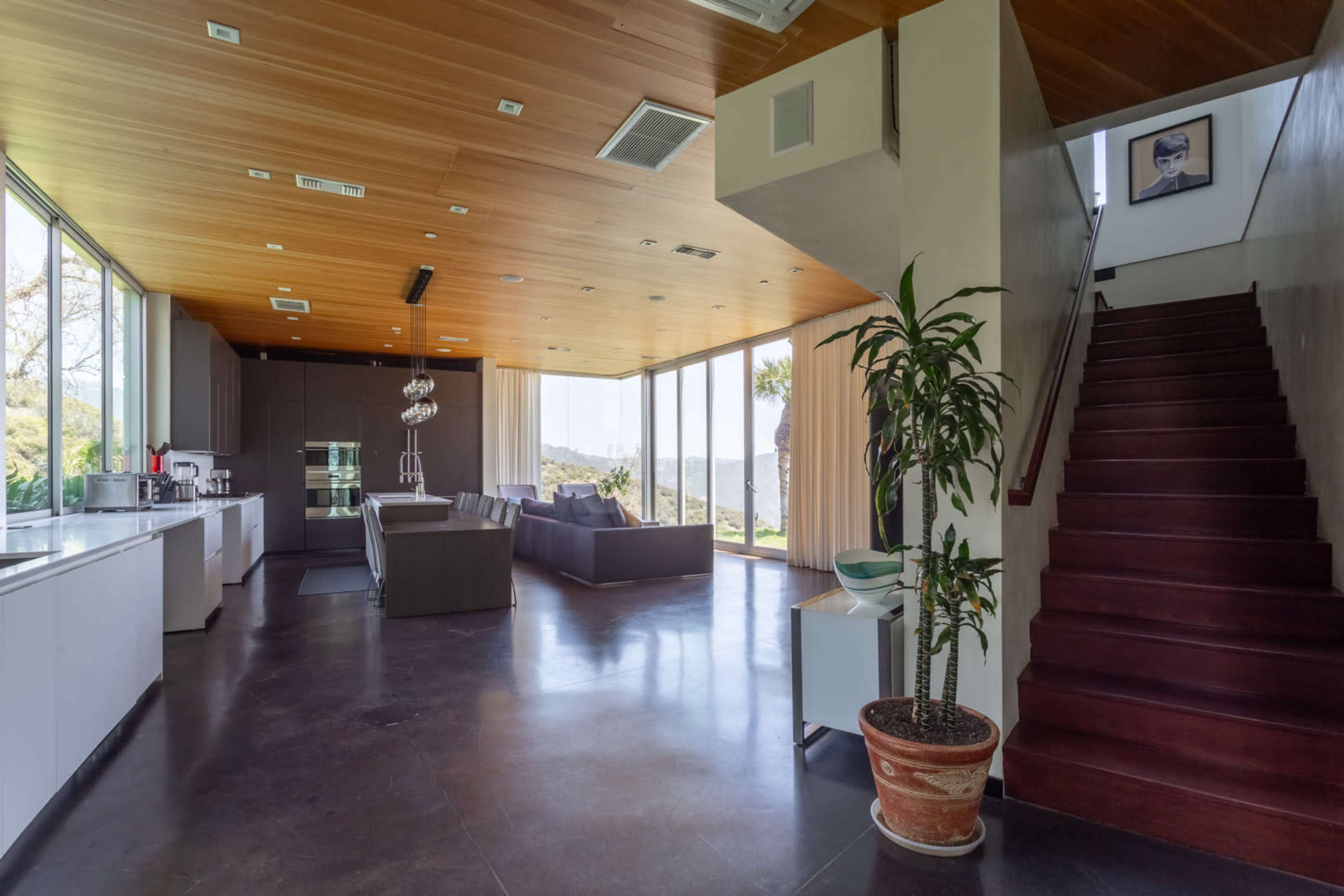 A modern open-plan living space with a kitchen, dining area, and staircase leading to an upper level, featuring large windows that provide natural light and a view of the outdoors.
