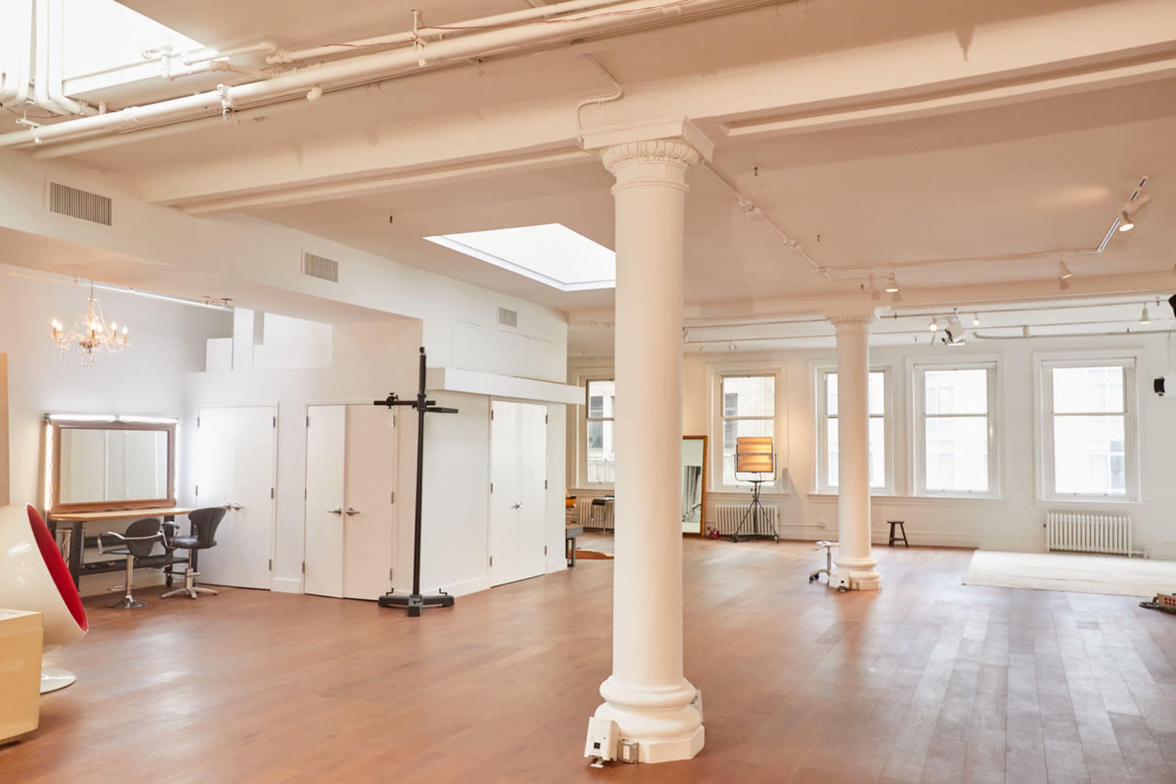 The image shows a spacious, well-lit room with wooden floors, large windows, and several white columns, featuring various furnishings and divided areas.