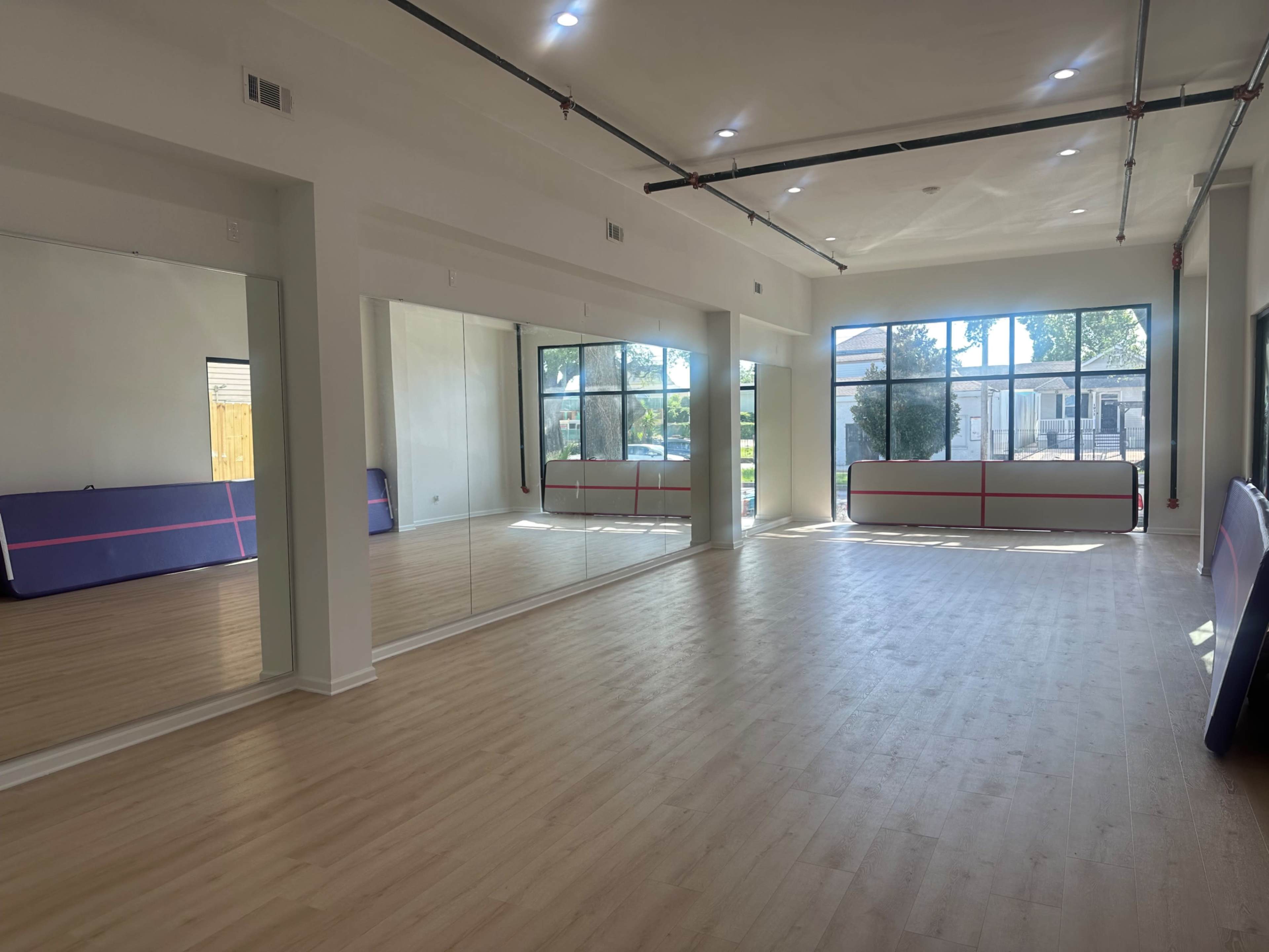The image shows an empty, well-lit room with large windows, a wooden floor, and mirrors along one wall.