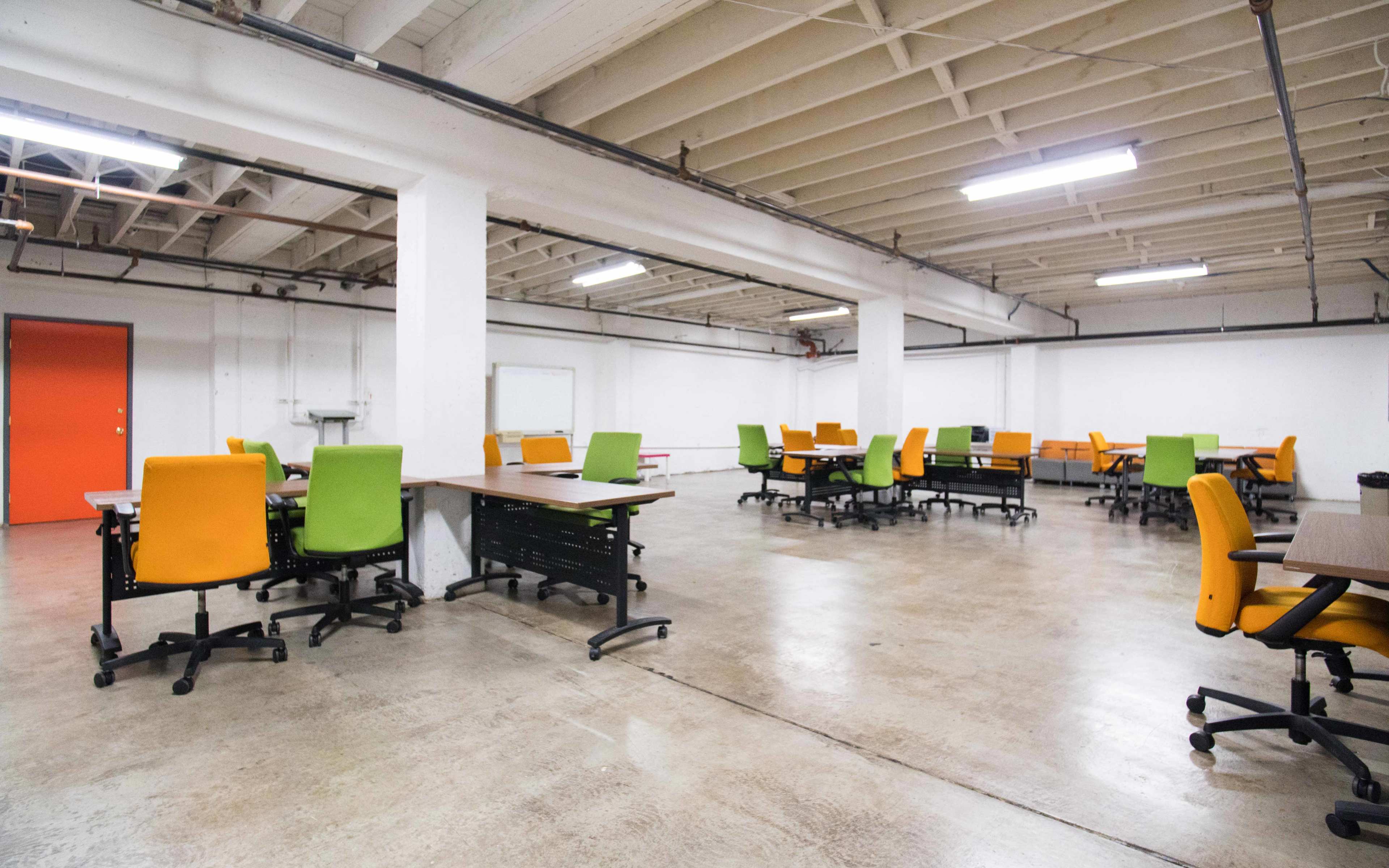 A spacious, minimally furnished office area features multiple desks with brightly colored chairs arranged in a grid pattern on a concrete floor.