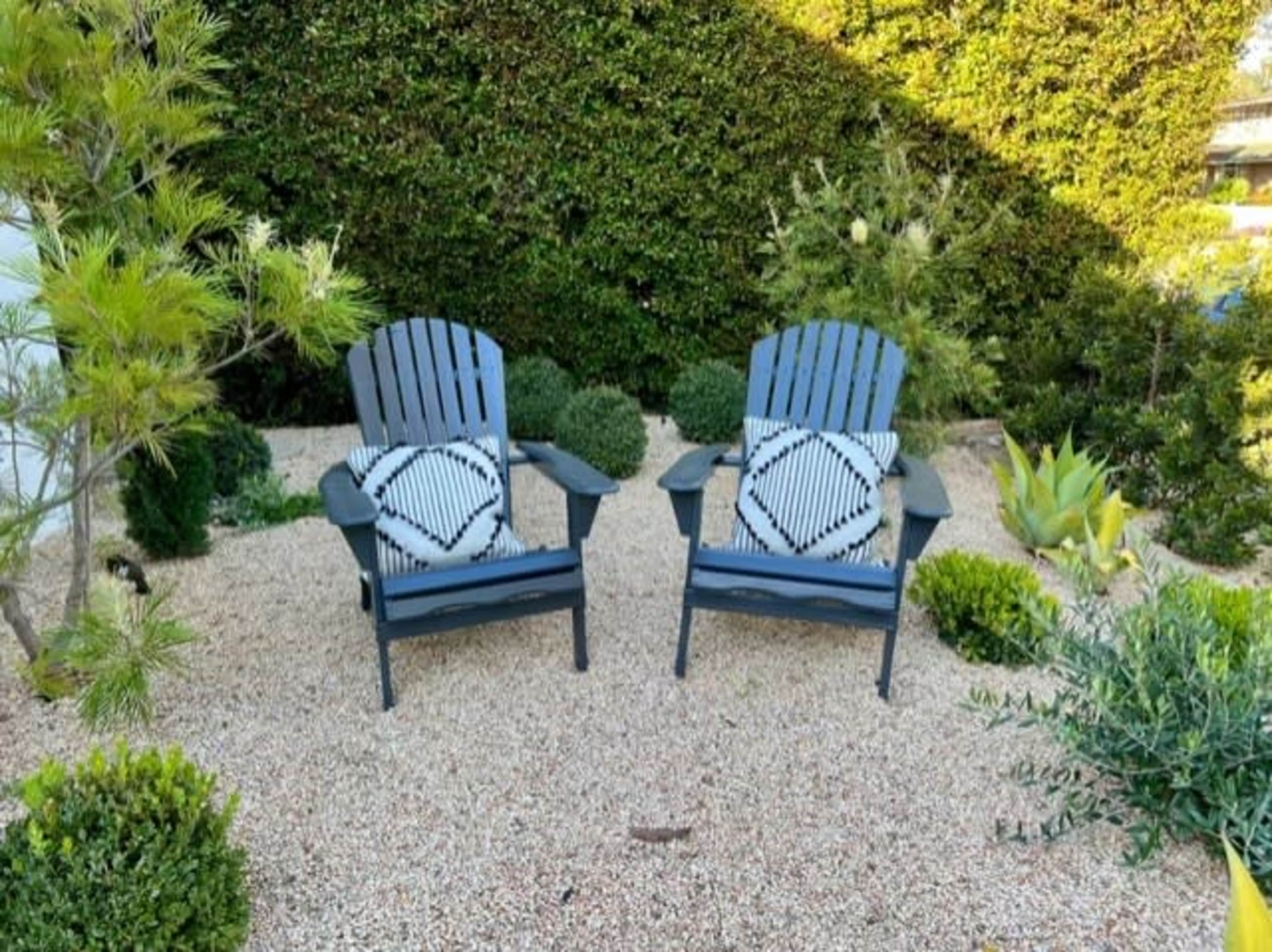 Two Adirondack chairs with patterned cushions are placed on a gravel surface surrounded by various greenery.
